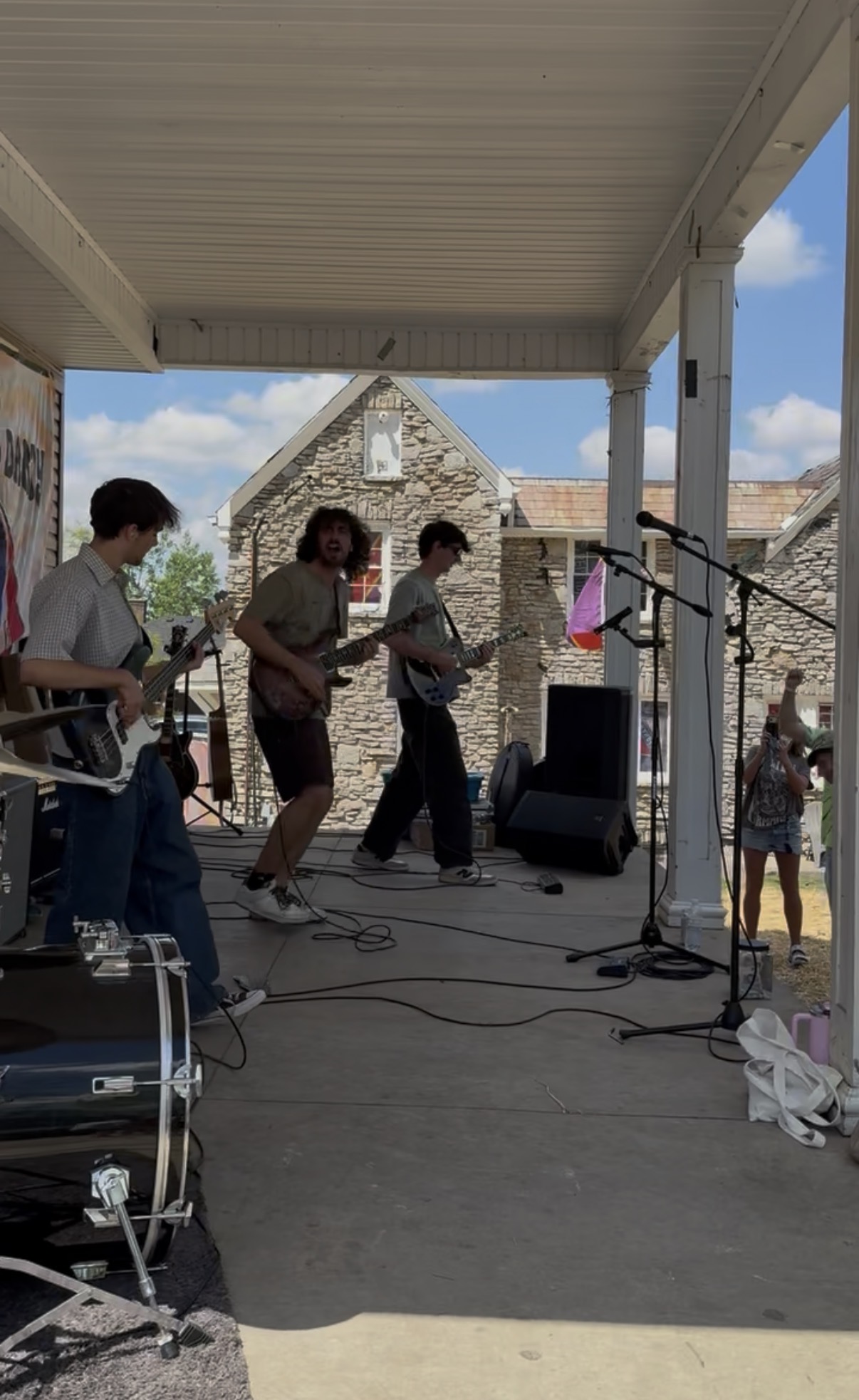 Group of people performing music on an outdoor stage with microphones and instruments, under a covered area, with a stone building and blue sky in background.