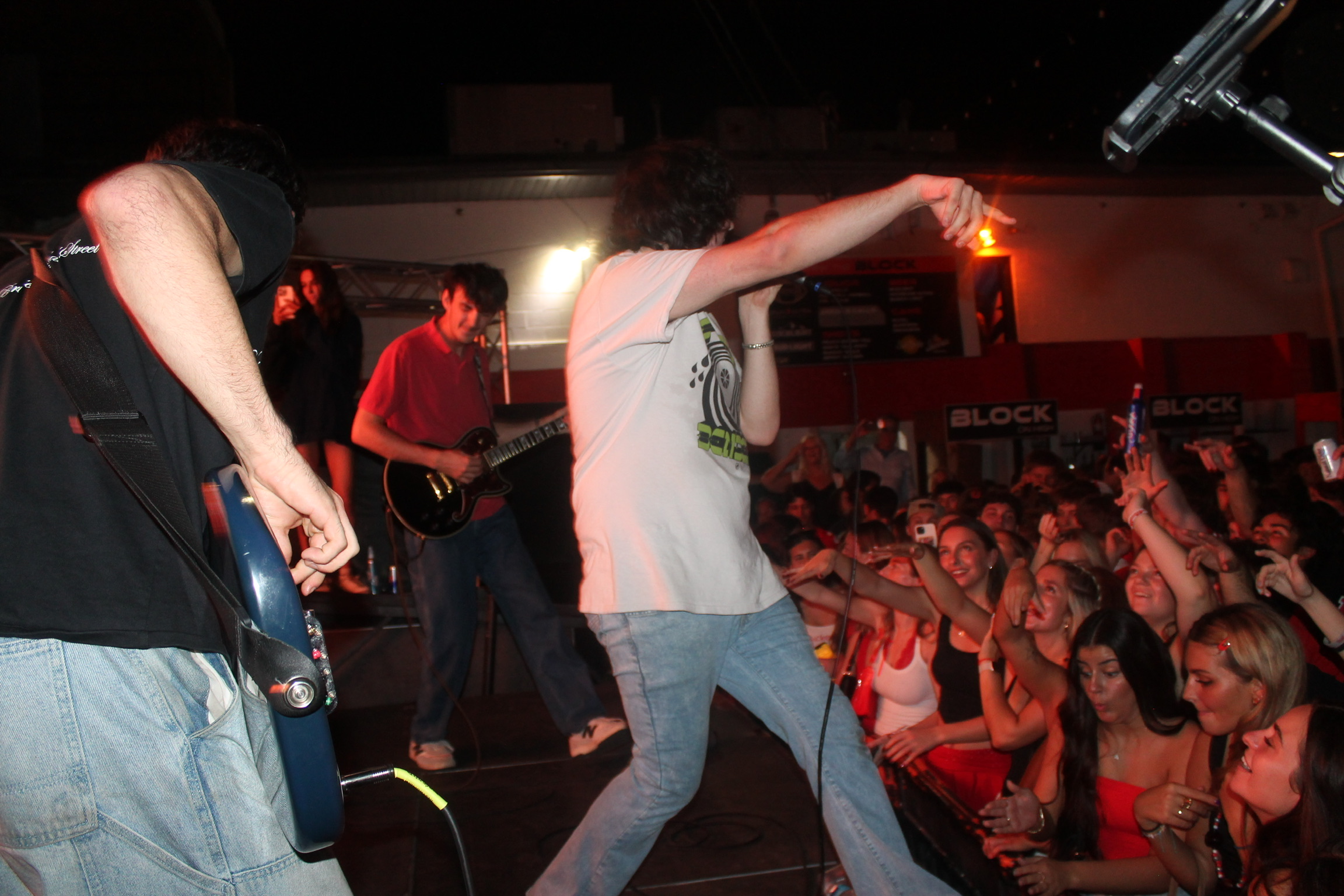 Band performs on stage with audience cheering and raising hands, red lighting in venue, people enjoying concert.