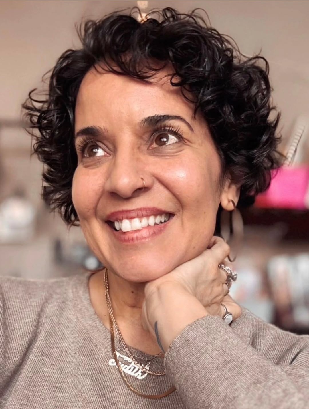 Woman with short curly hair smiling, resting her chin on her hand, wearing a gray sweater and jewelry.