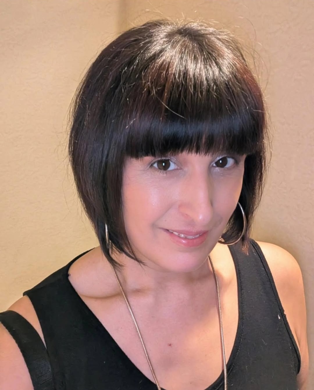 Young woman with short dark hair and bangs, smiling, wearing a black sleeveless top and a necklace.