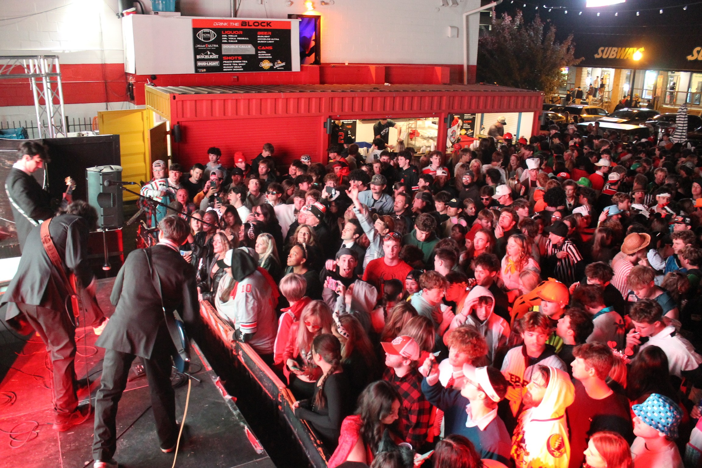 Large crowd of people gathered outside a building with red and yellow exterior, some waiting in line, at night.
