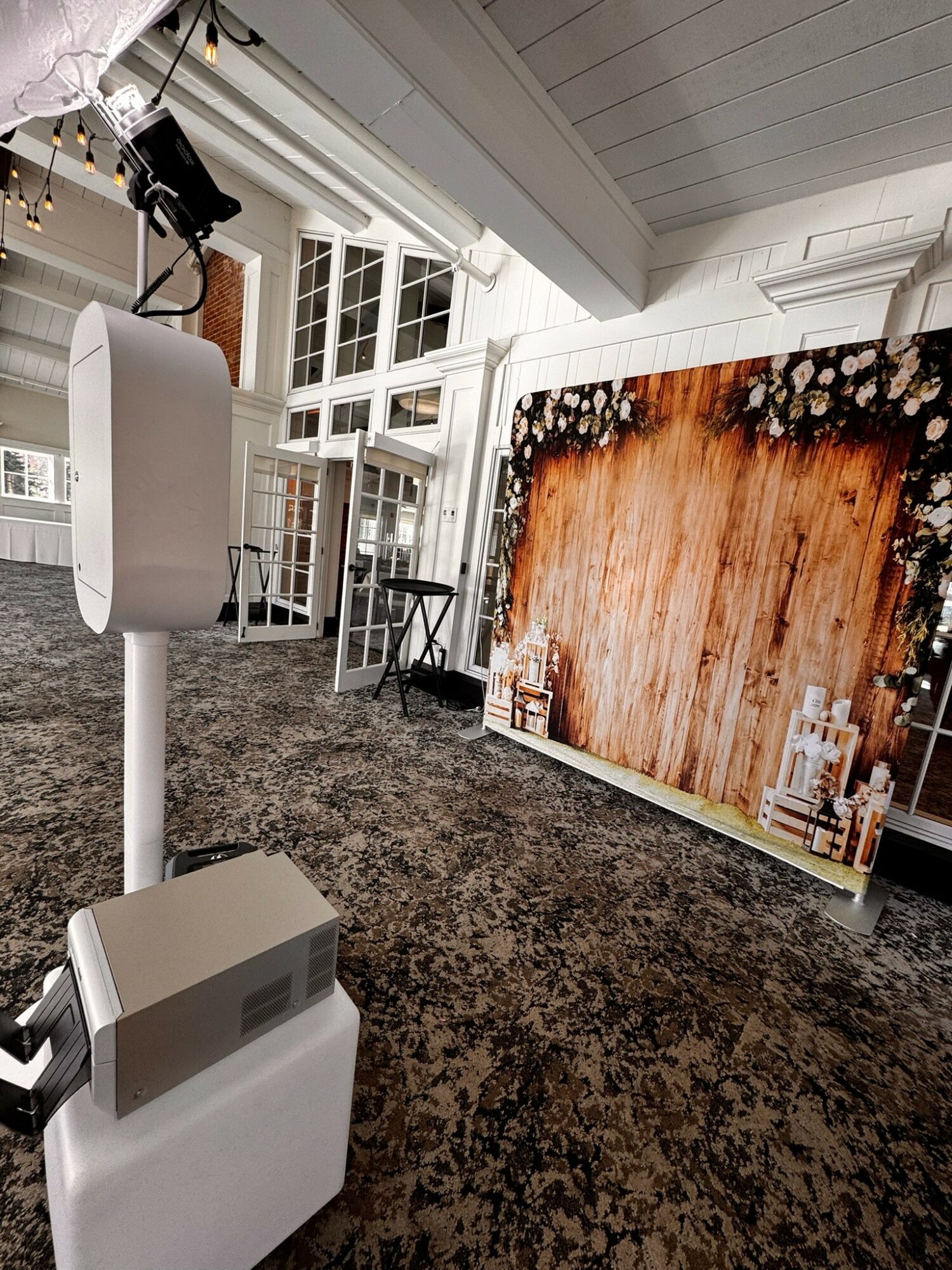 Indoor space with a large wooden backdrop, white walls, and a windowed ceiling, featuring a speaker on a stand and a carpeted floor.