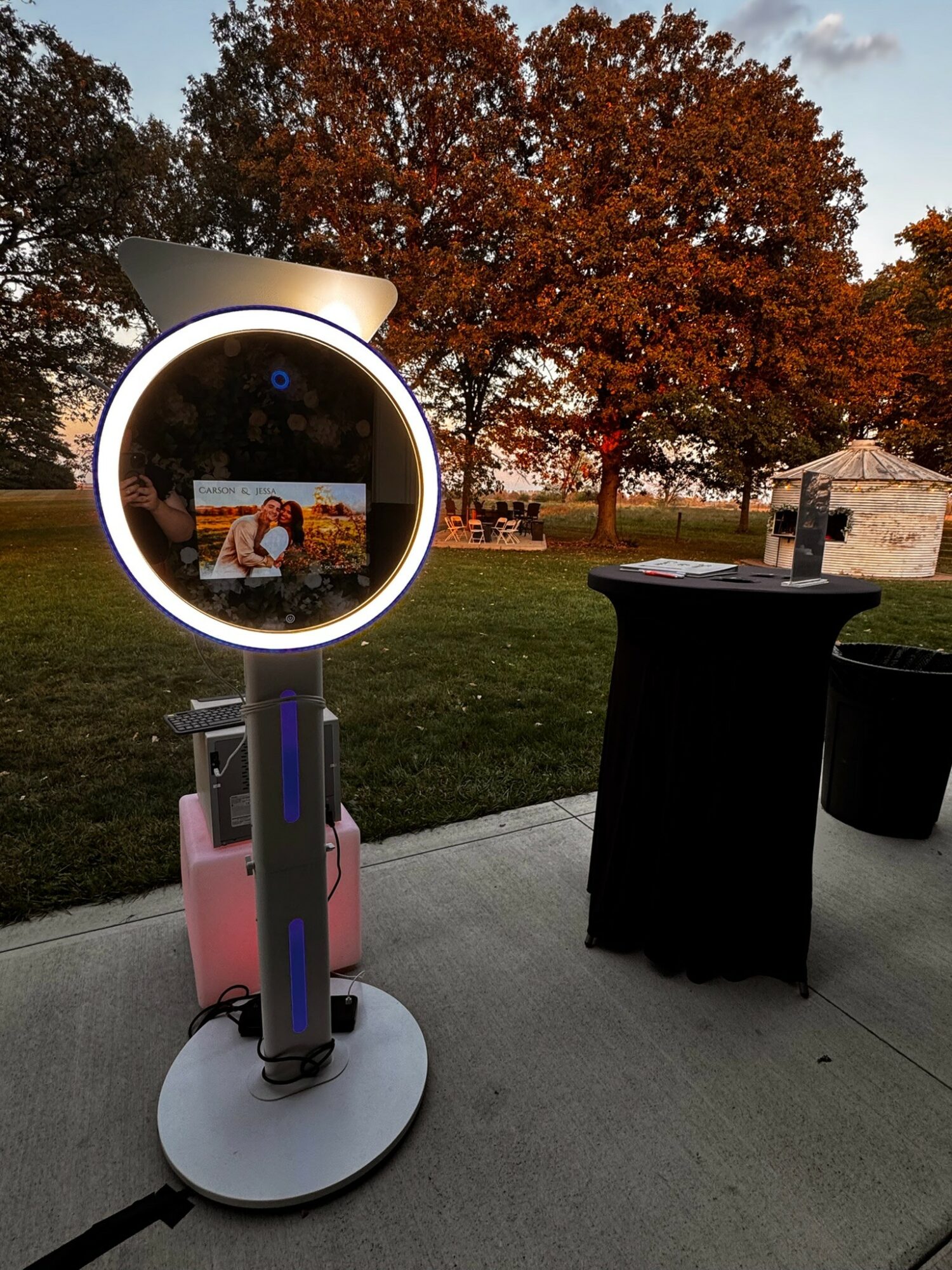 Photo of a photo booth with a circular light, outdoors with trees and a person in the reflection, during autumn.