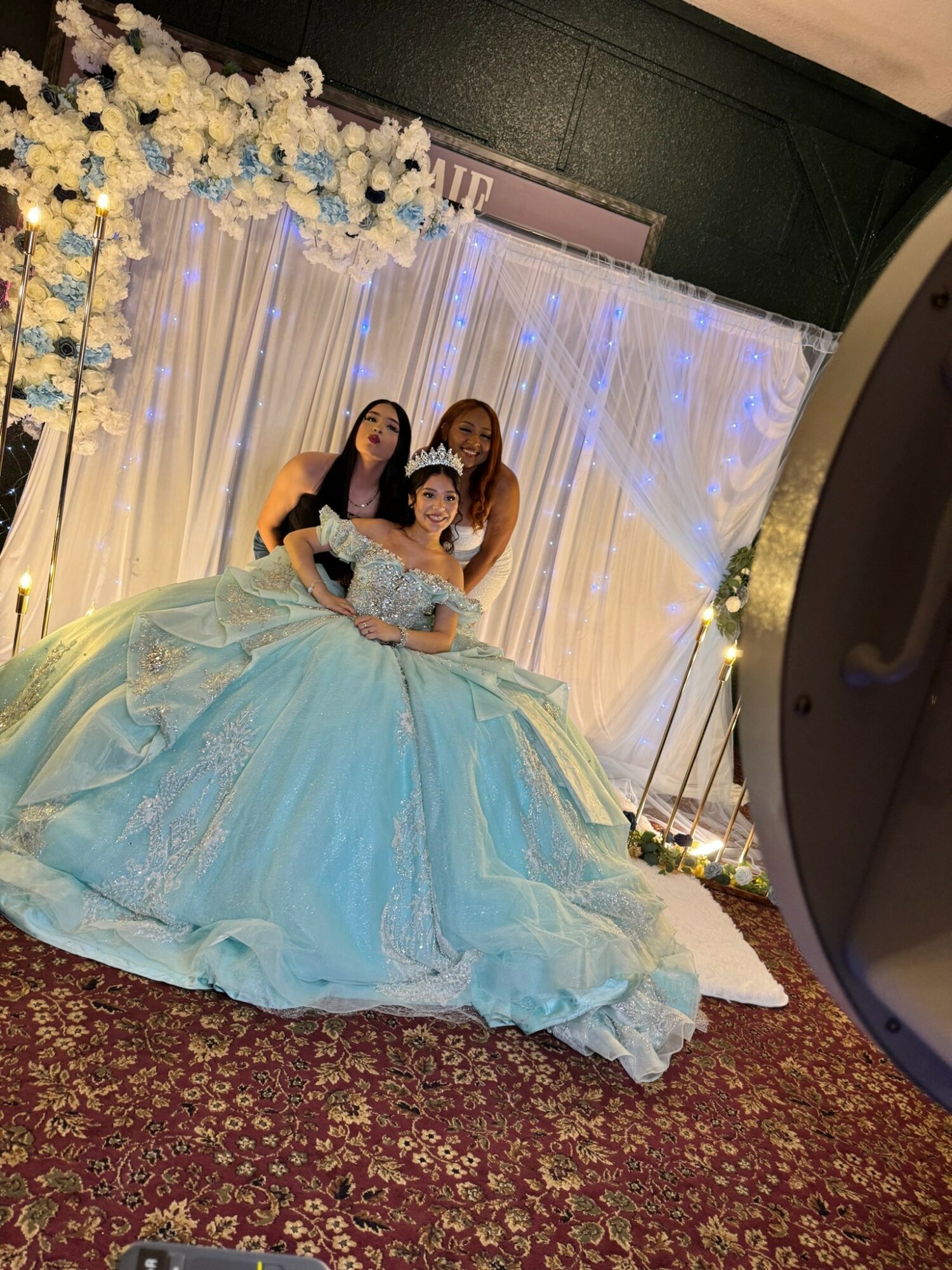 Three people, one in a blue gown, pose in front of a decorated backdrop with flowers and lights.