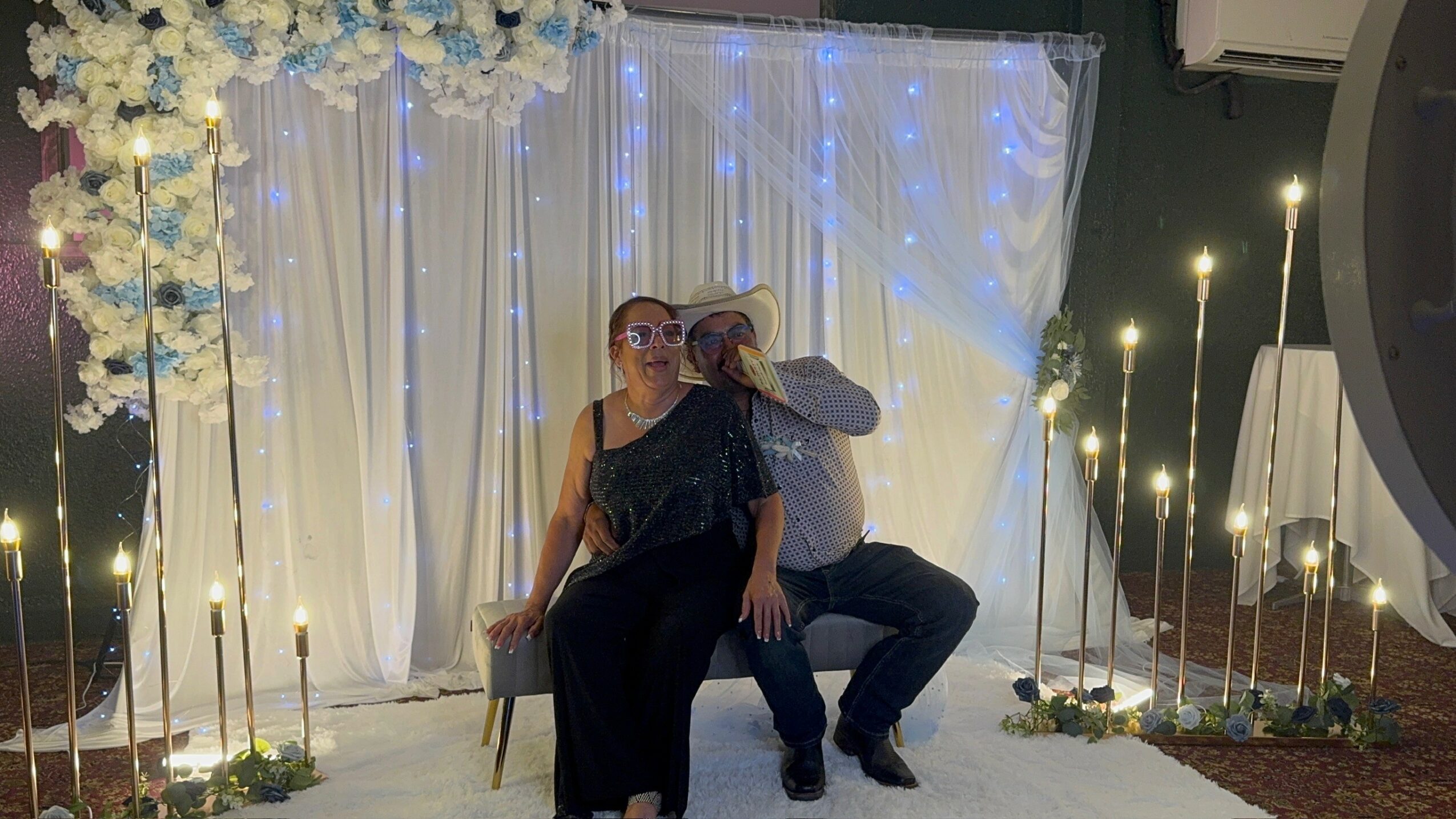 Two women sitting in front of a decorated backdrop with white curtains and string lights, one wearing sunglasses.