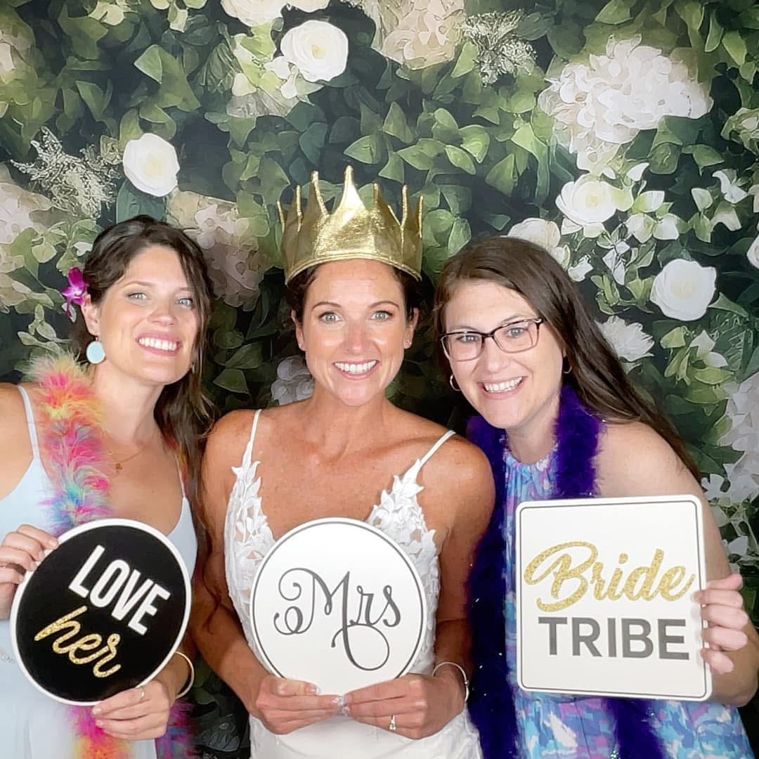 Three women smiling at a celebration, one wearing a crown, holding signs that say 'LOVE her,' 'Mrs,' and 'Bride TRIBE,' with a floral background.