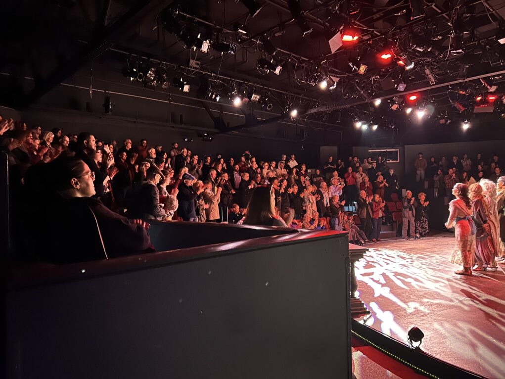 Audience watching a performance on stage in a dark theater with red lighting, some people standing and others seated.