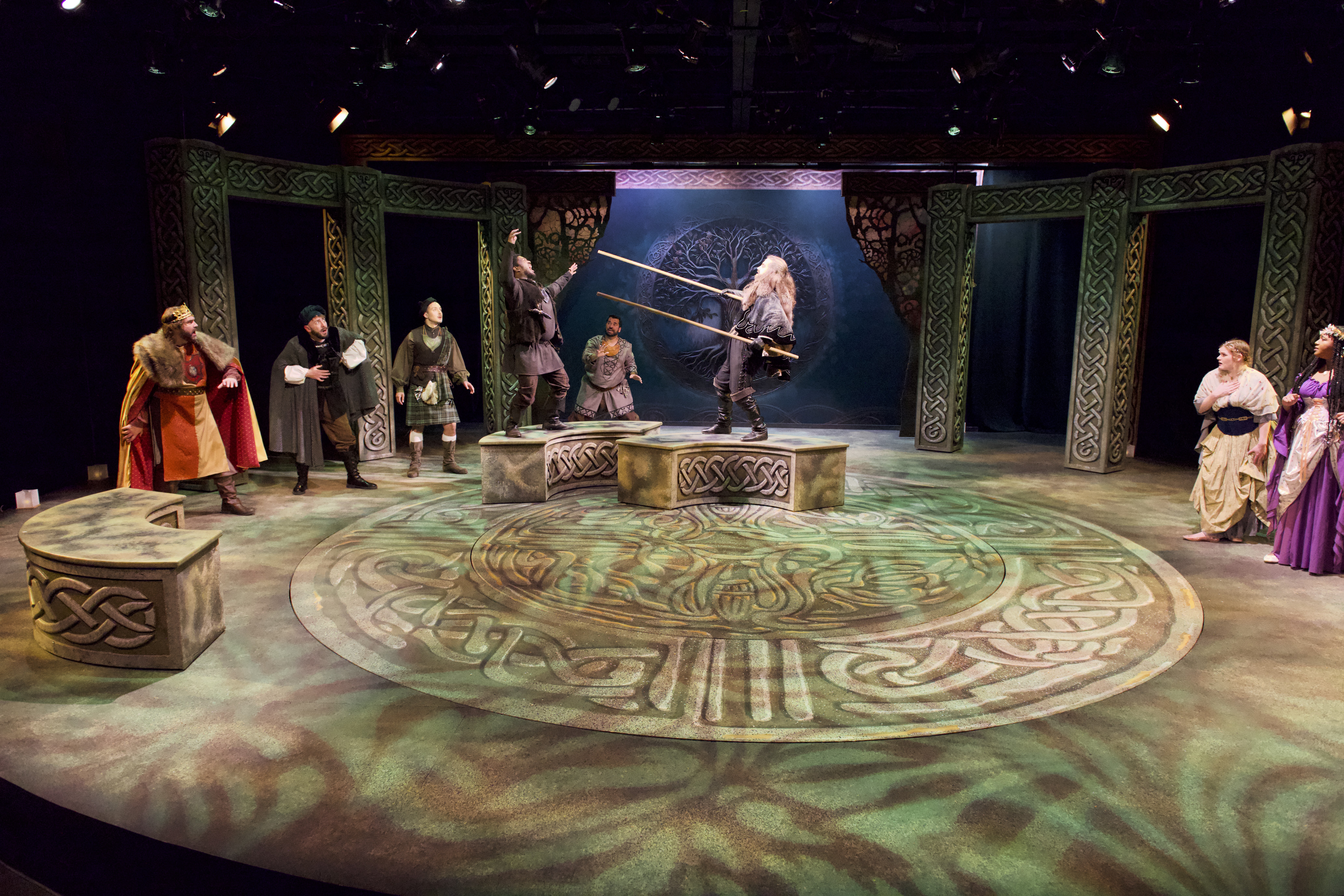 Theater stage with actors in period costumes, a person on a raised platform, and decorative set pieces, with a circular patterned floor.