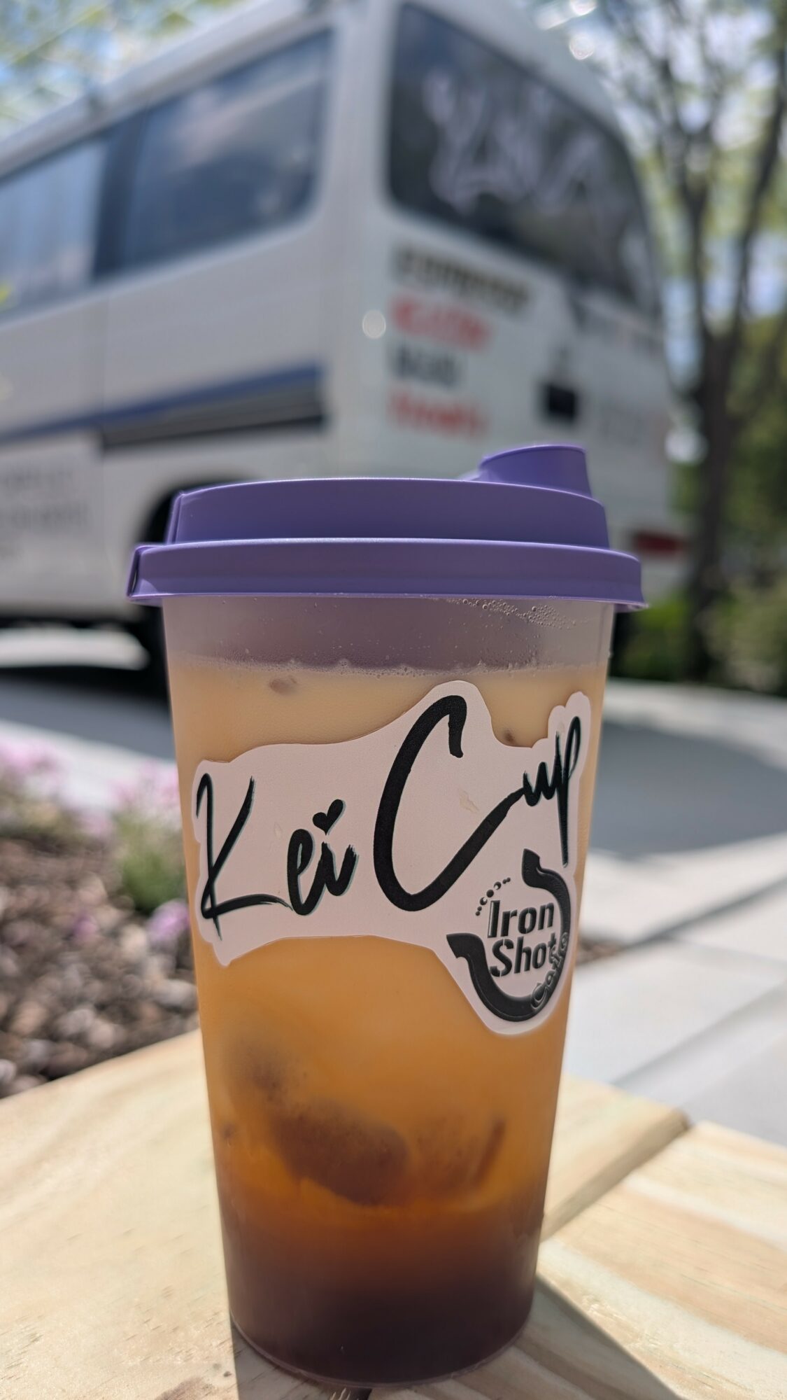 Iced coffee in a plastic cup with a purple lid, placed outdoors with a bus in the background.