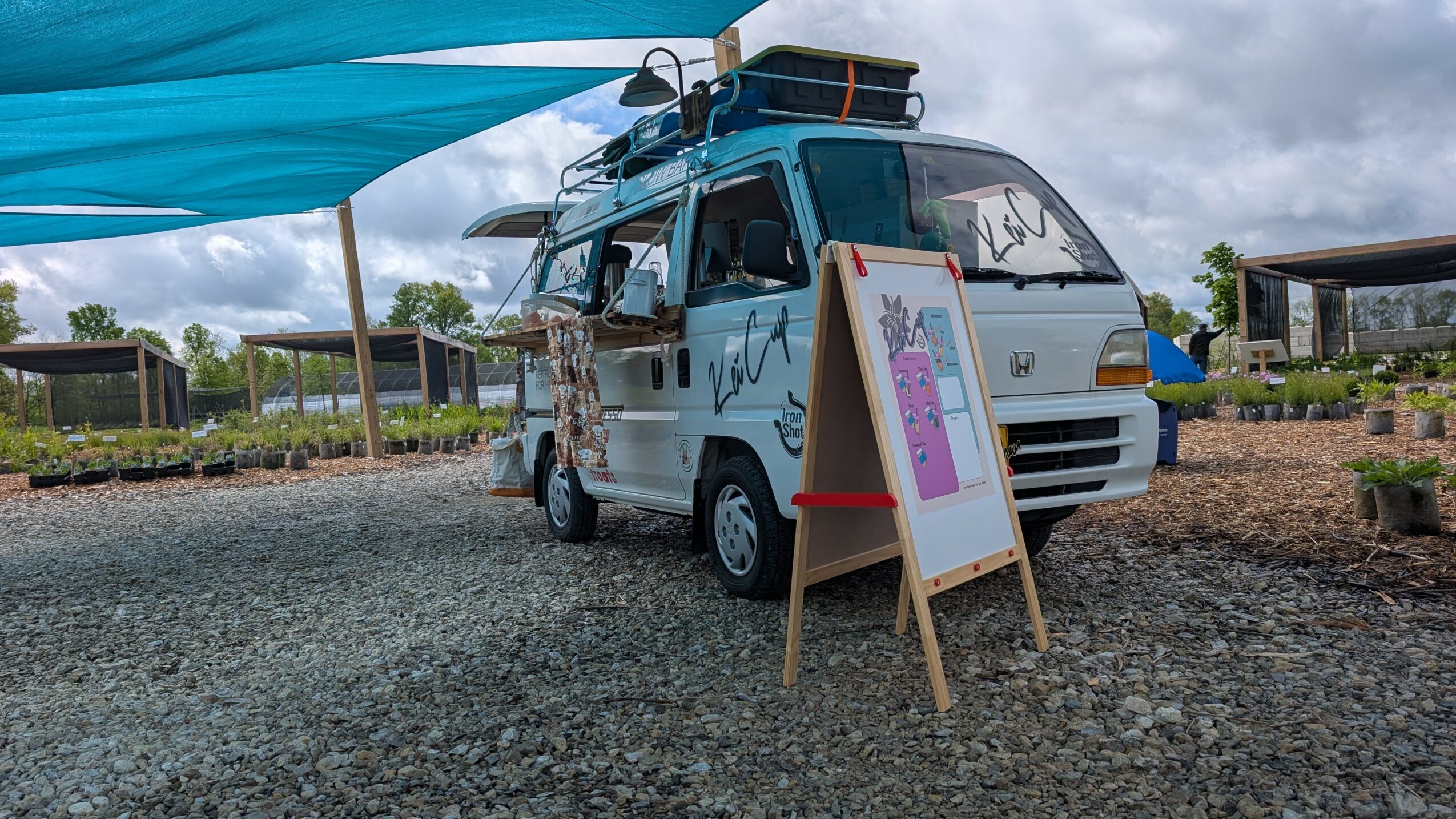 Small van with equipment on top, parked outdoors with a signboard nearby, under a blue canopy, on gravel ground.
