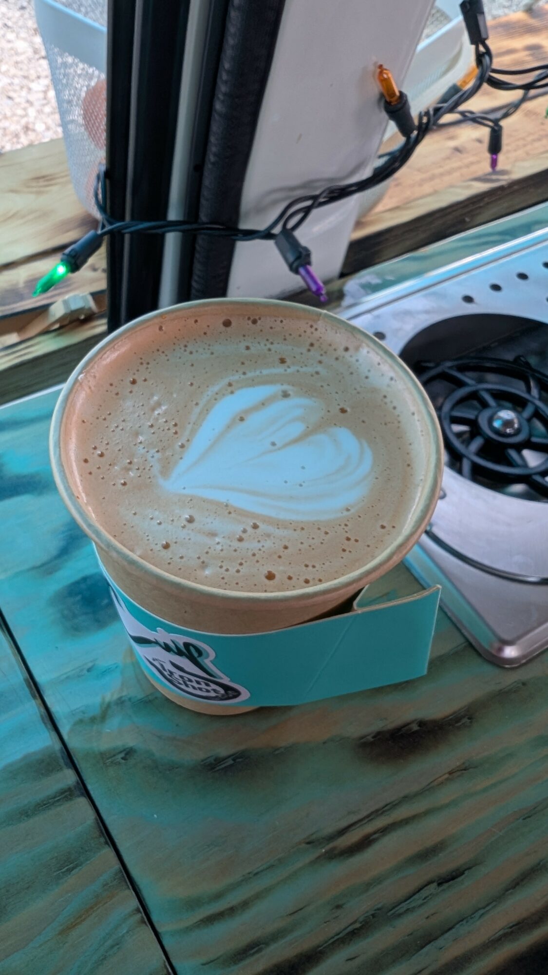 Cup of coffee with heart-shaped foam art on top, placed on a wooden surface near a window with wires and a stove in the background.