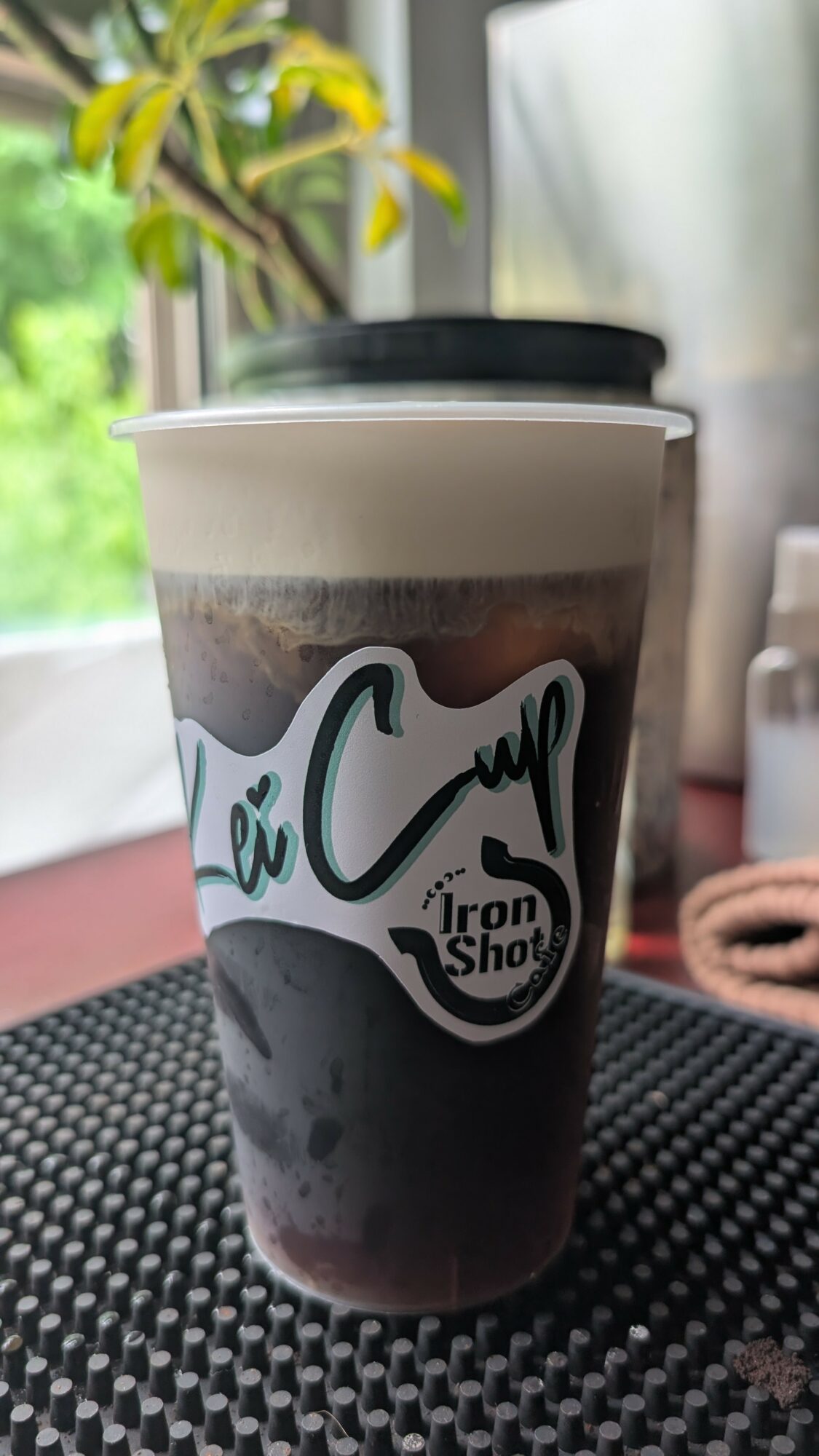 A coffee cup with a logo, placed on a textured surface near a window with greenery outside.