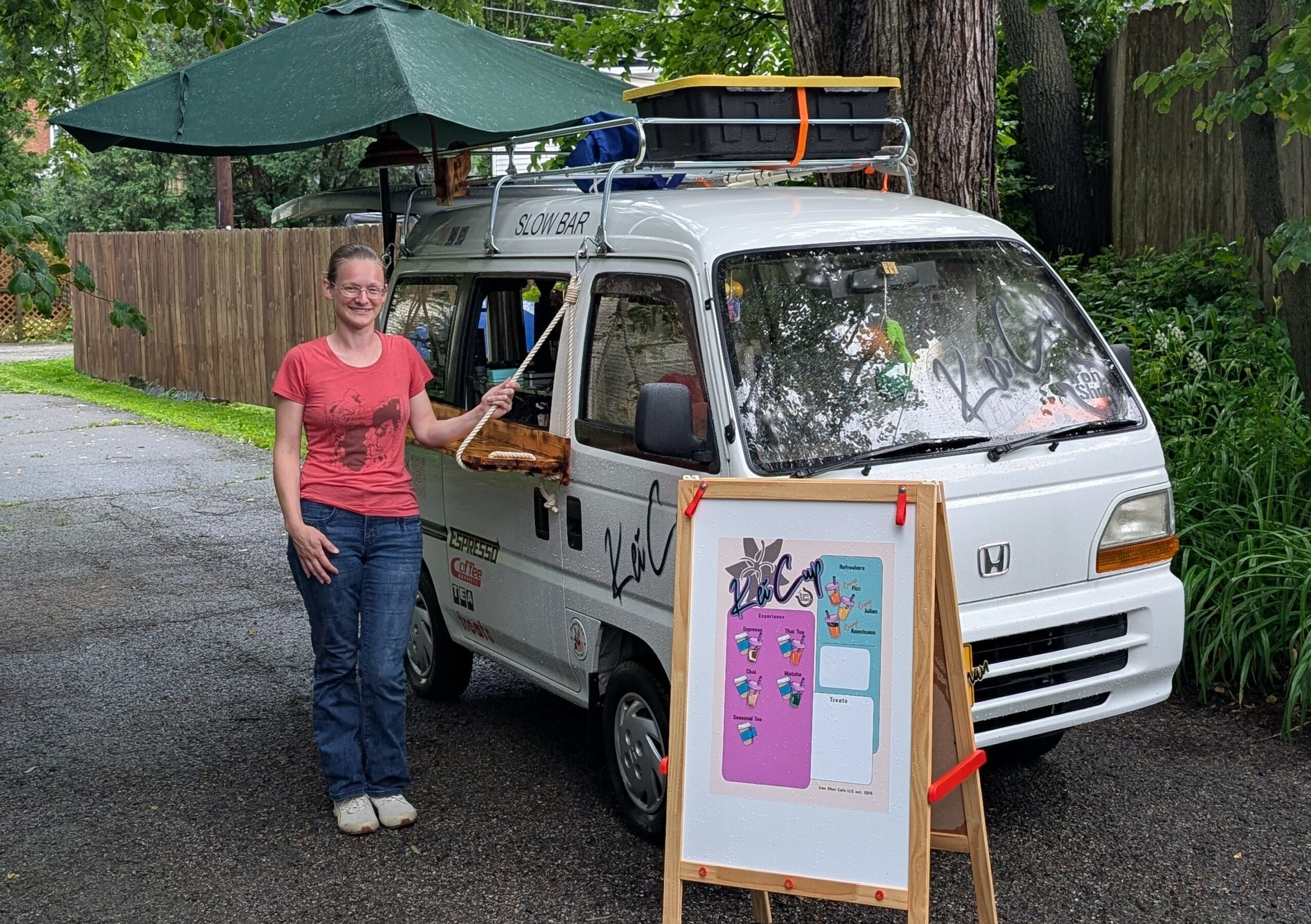 Woman standing next to a small white van with a pink and blue display board outside, trees and a wooden fence in background.