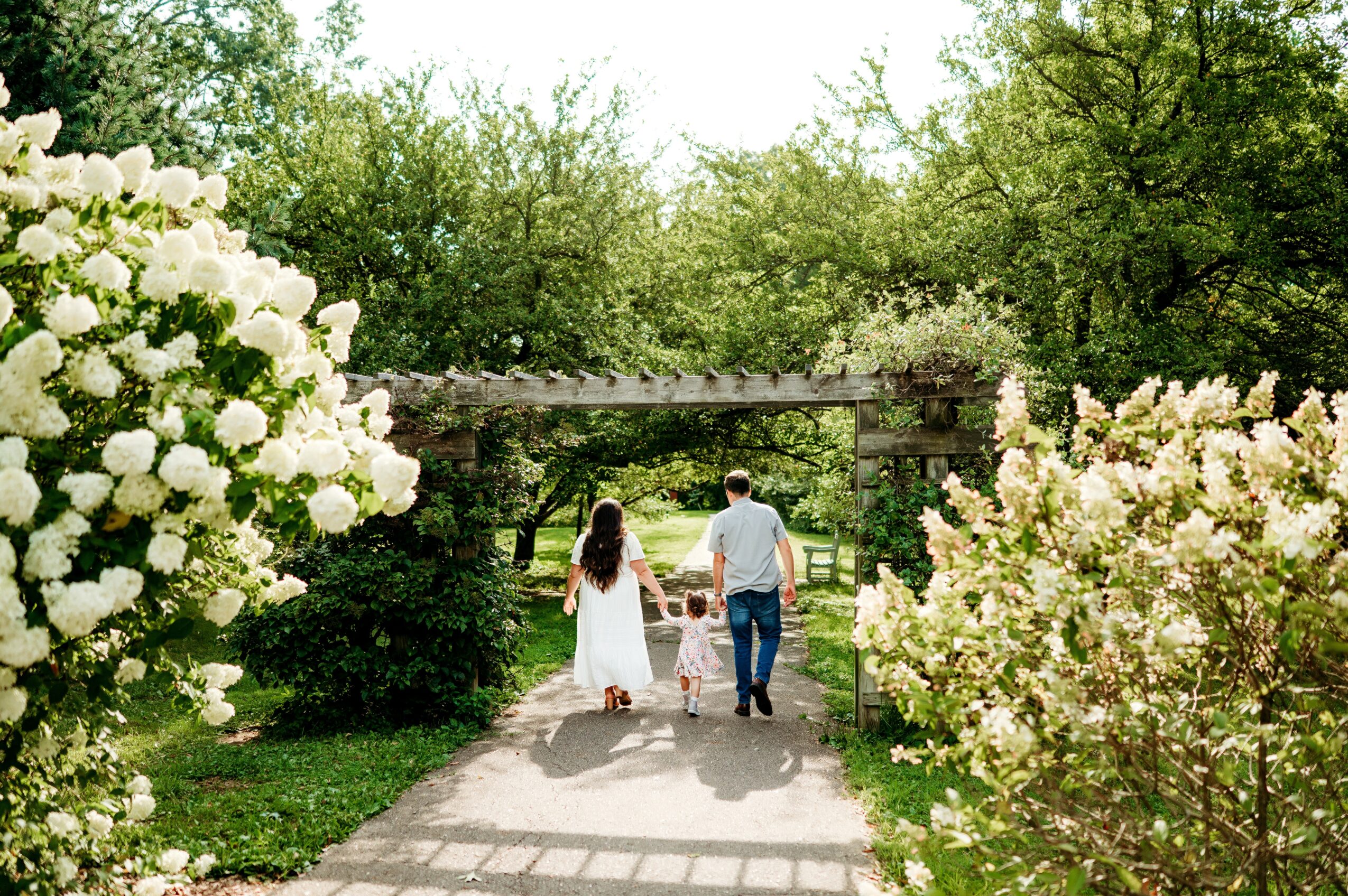 A family walking on a path surrounded by blooming bushes and trees, holding hands, with a bridge in the background.