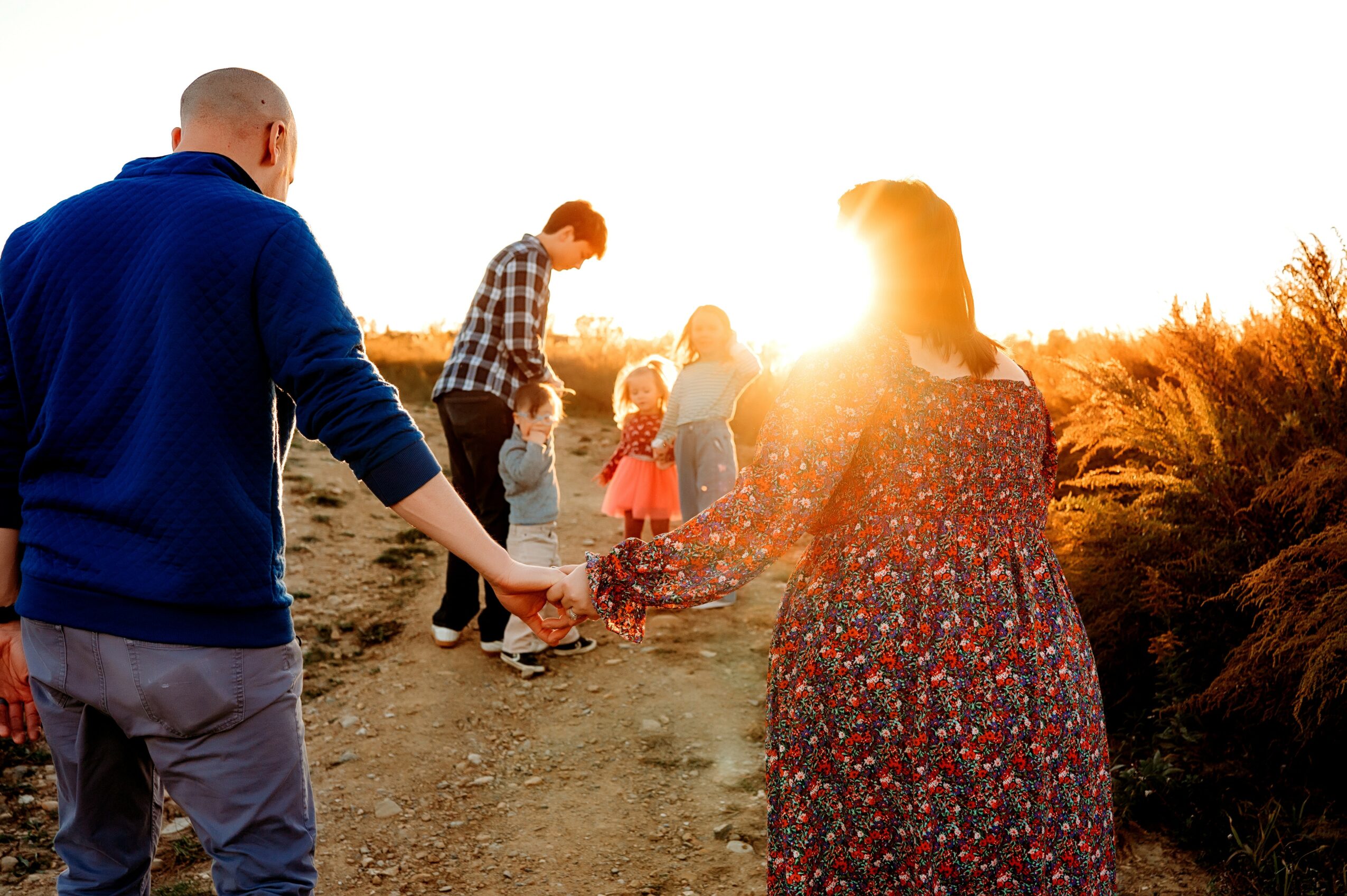 Group of people holding hands outdoors during sunset, walking along a dirt path with trees and bushes nearby.