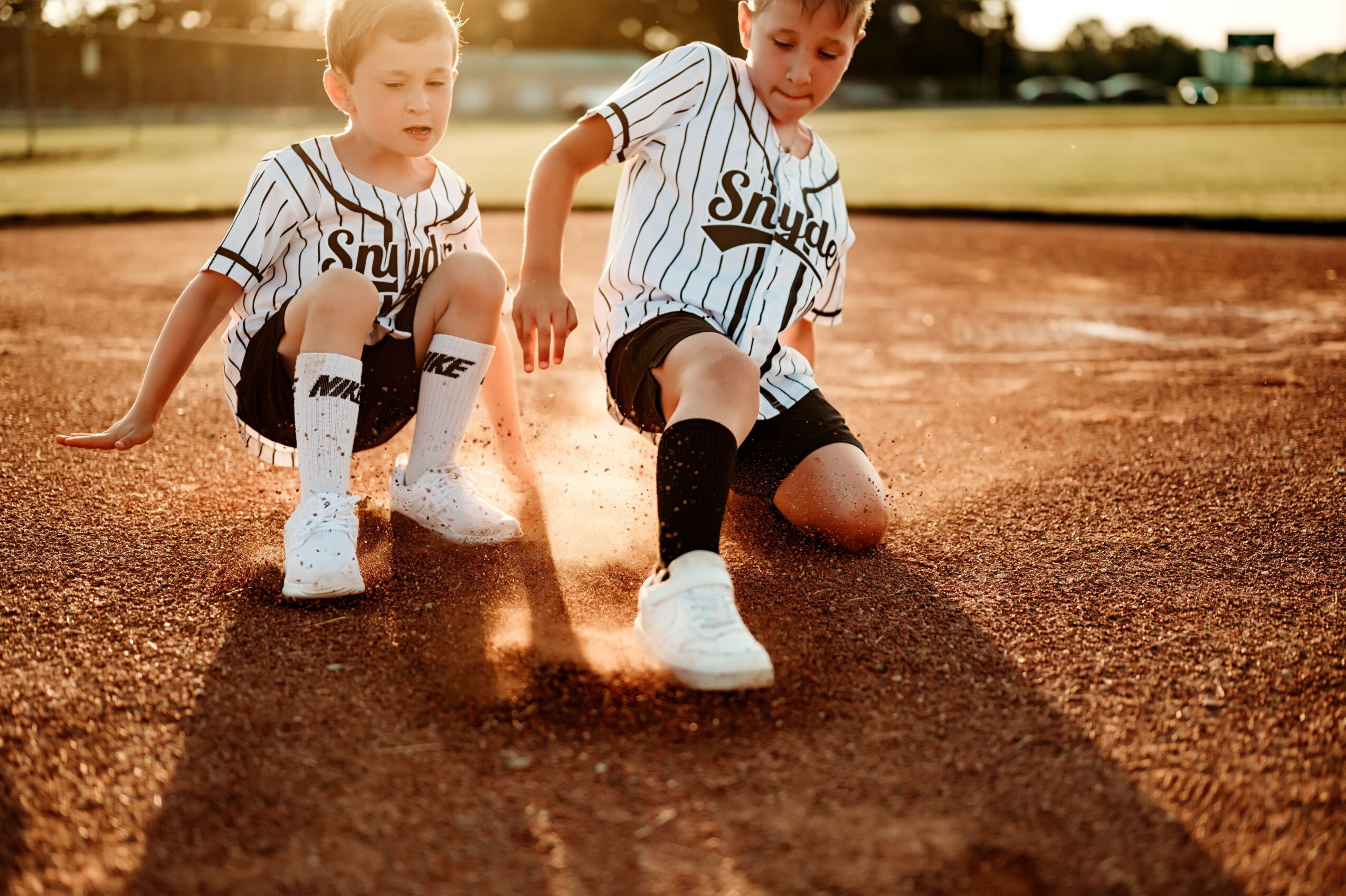 Two children in baseball uniforms kneel on a baseball field, touching the ground with their hands, with a blurred background.
