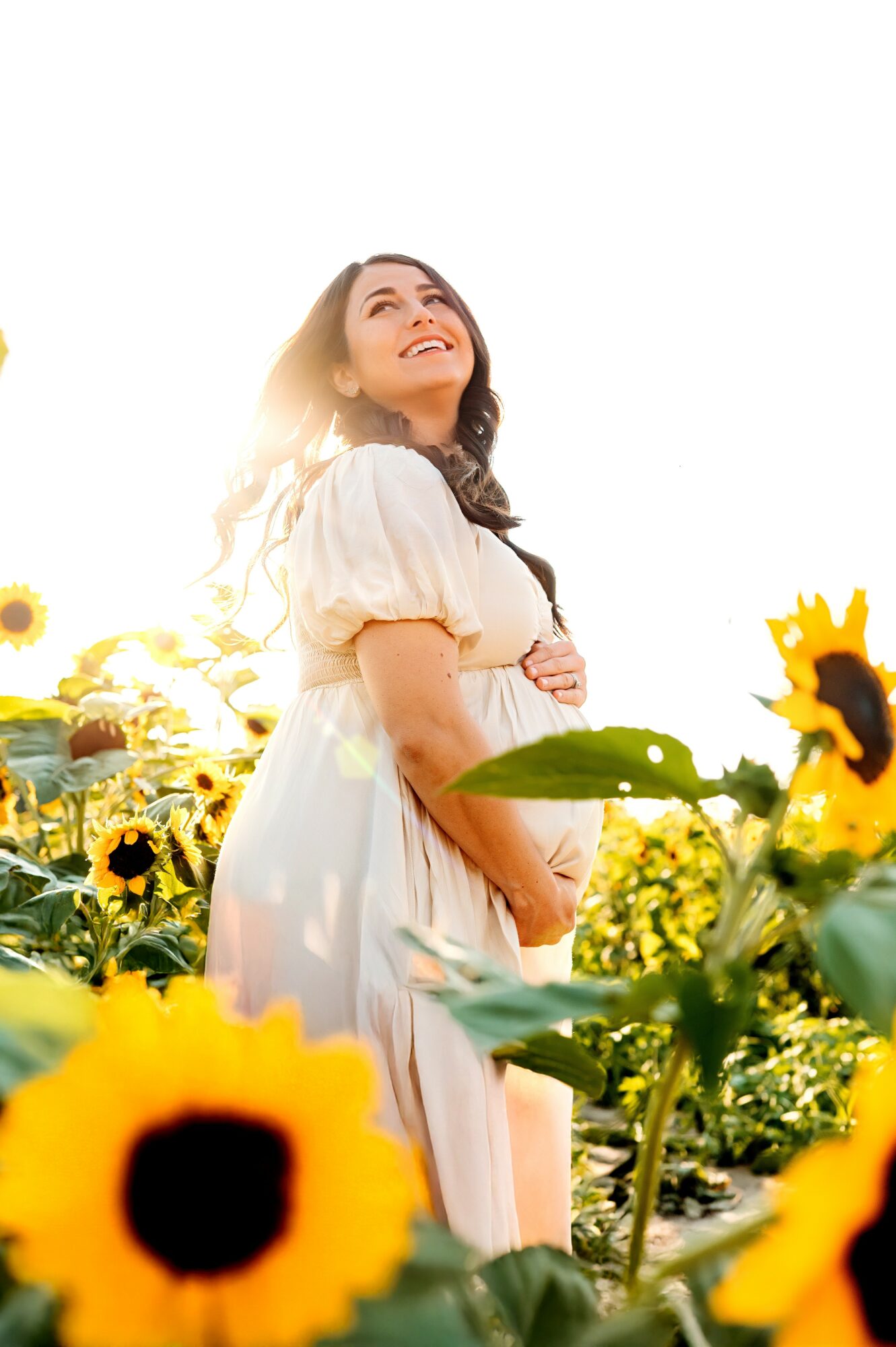 Woman in white dress standing among sunflowers, smiling and looking upward, sunlight behind her.