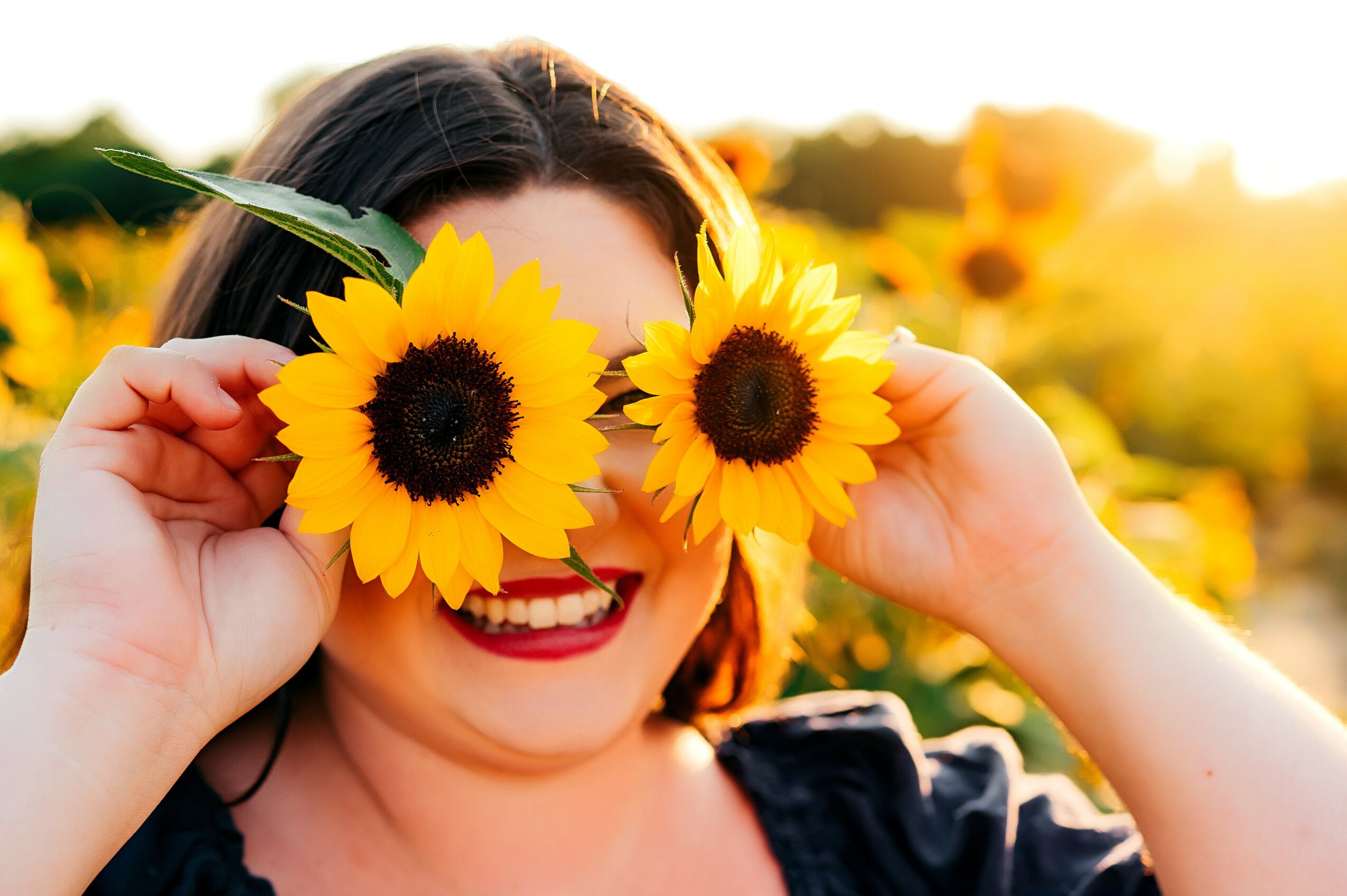 Person holding two sunflowers in front of their eyes, smiling in a sunflower field at sunset.