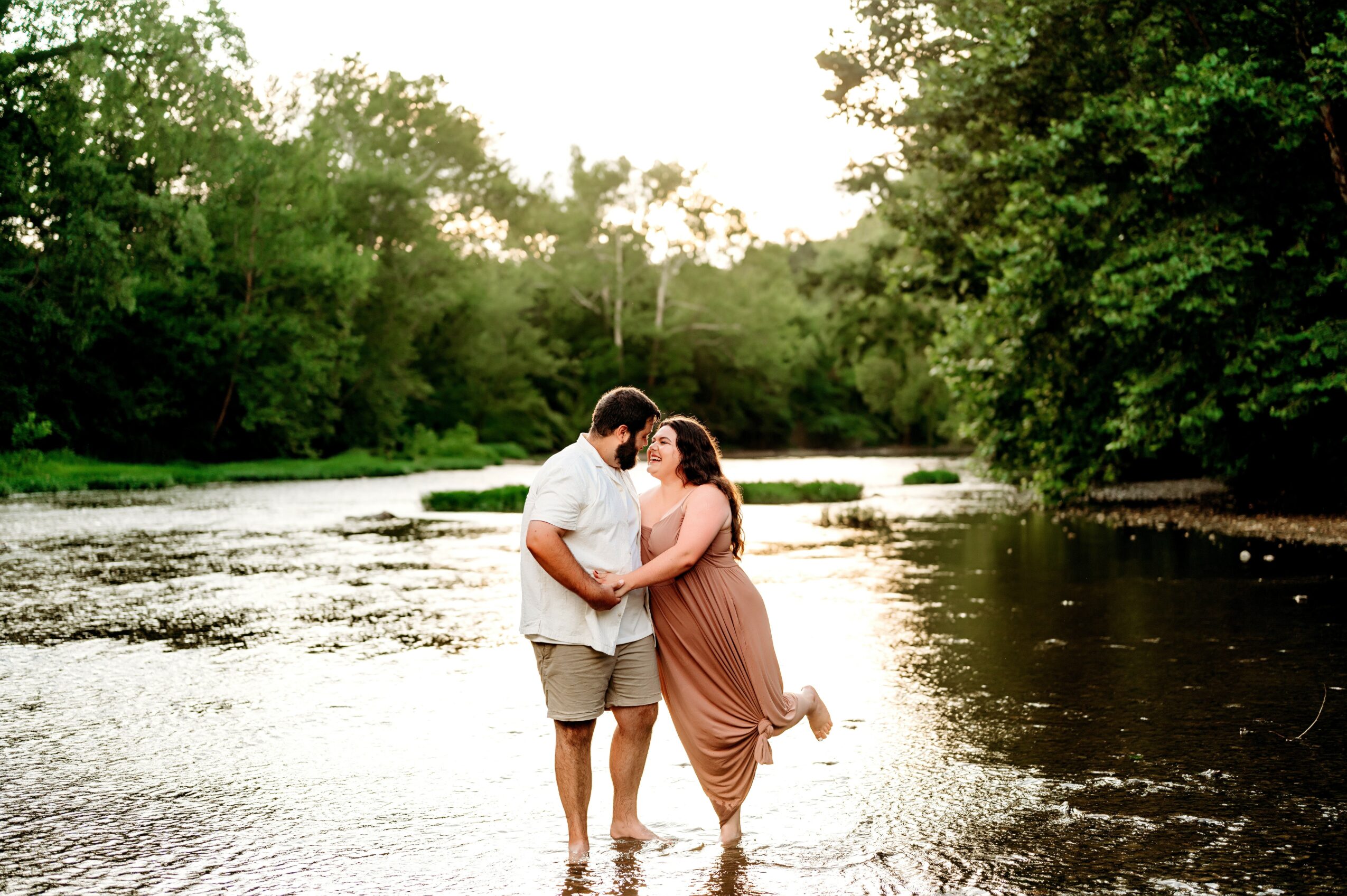A couple standing in a river, embracing, surrounded by trees and greenery, with sunlight filtering through the leaves.