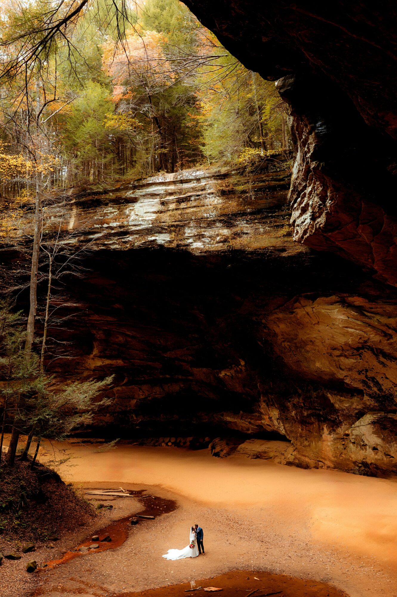 Two people stand on a sandy area inside a large cave with layered rock formations and trees outside visible at the entrance.