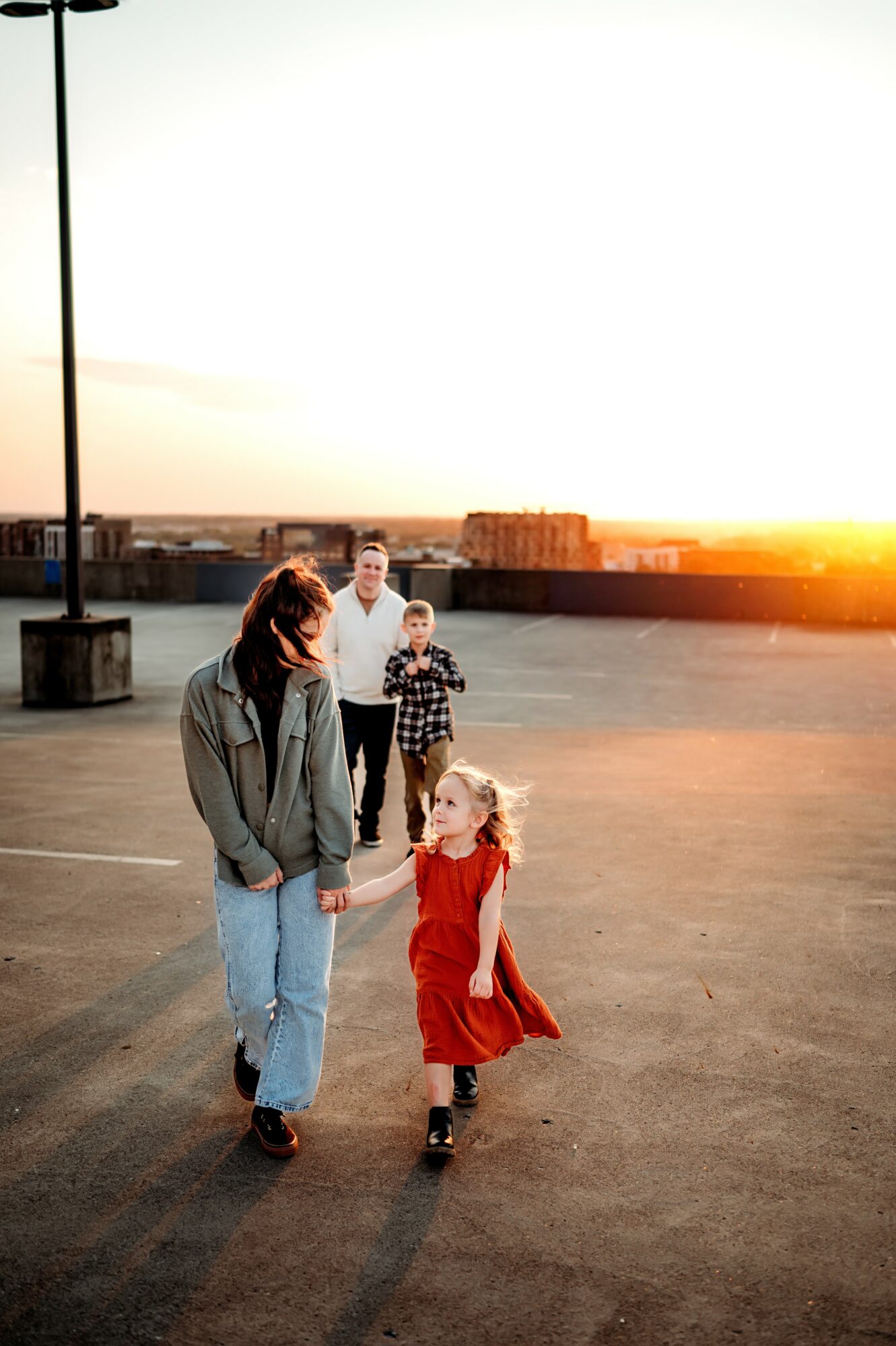 A woman and a young girl walk hand in hand in an open parking lot during sunset, with two people in the background.