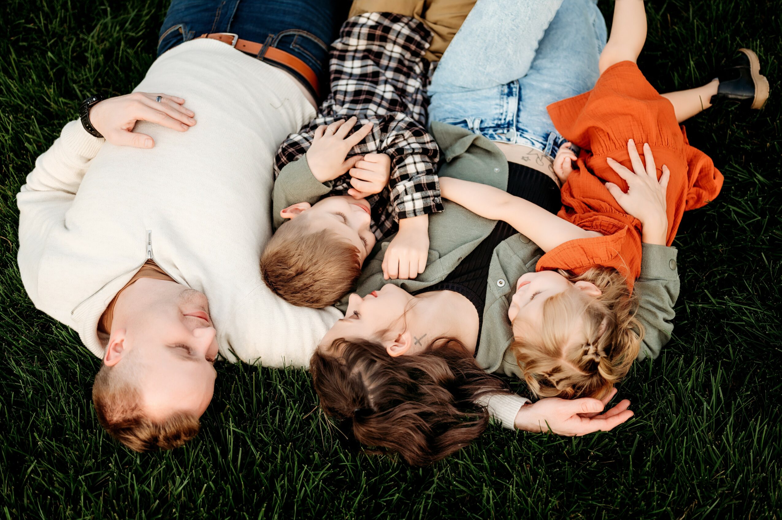 Four people lying on grass, two adults and two children, with heads close together, smiling and relaxing outdoors.