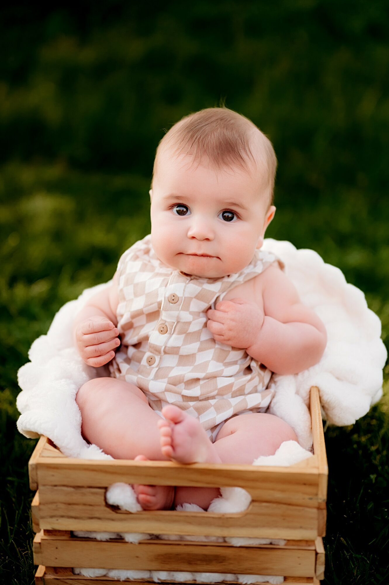 Baby sitting in a wooden box outdoors, wearing a checkered outfit, with a neutral expression, green background.