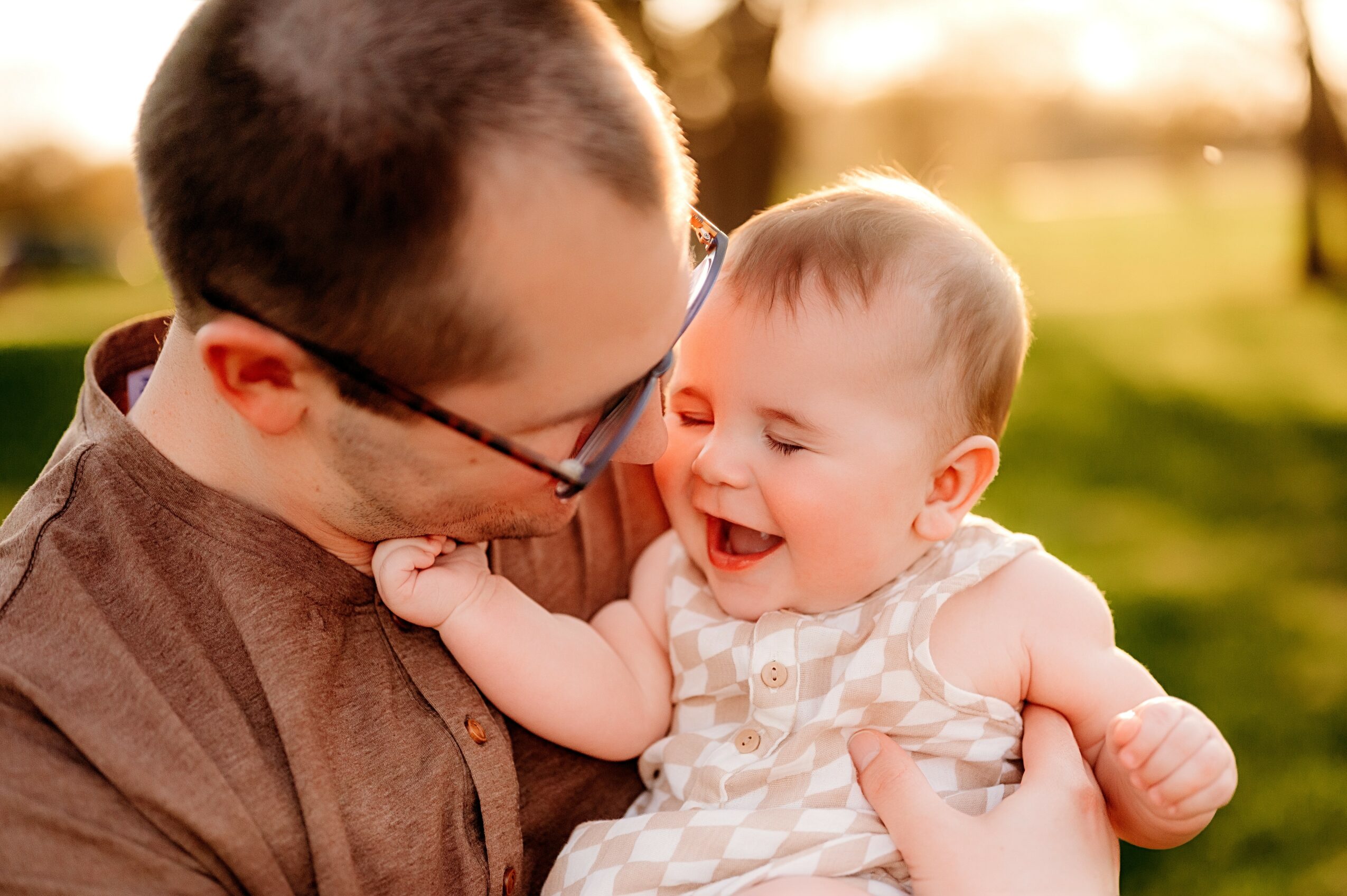 Adult holding a smiling baby outdoors during sunset, both looking at each other happily.
