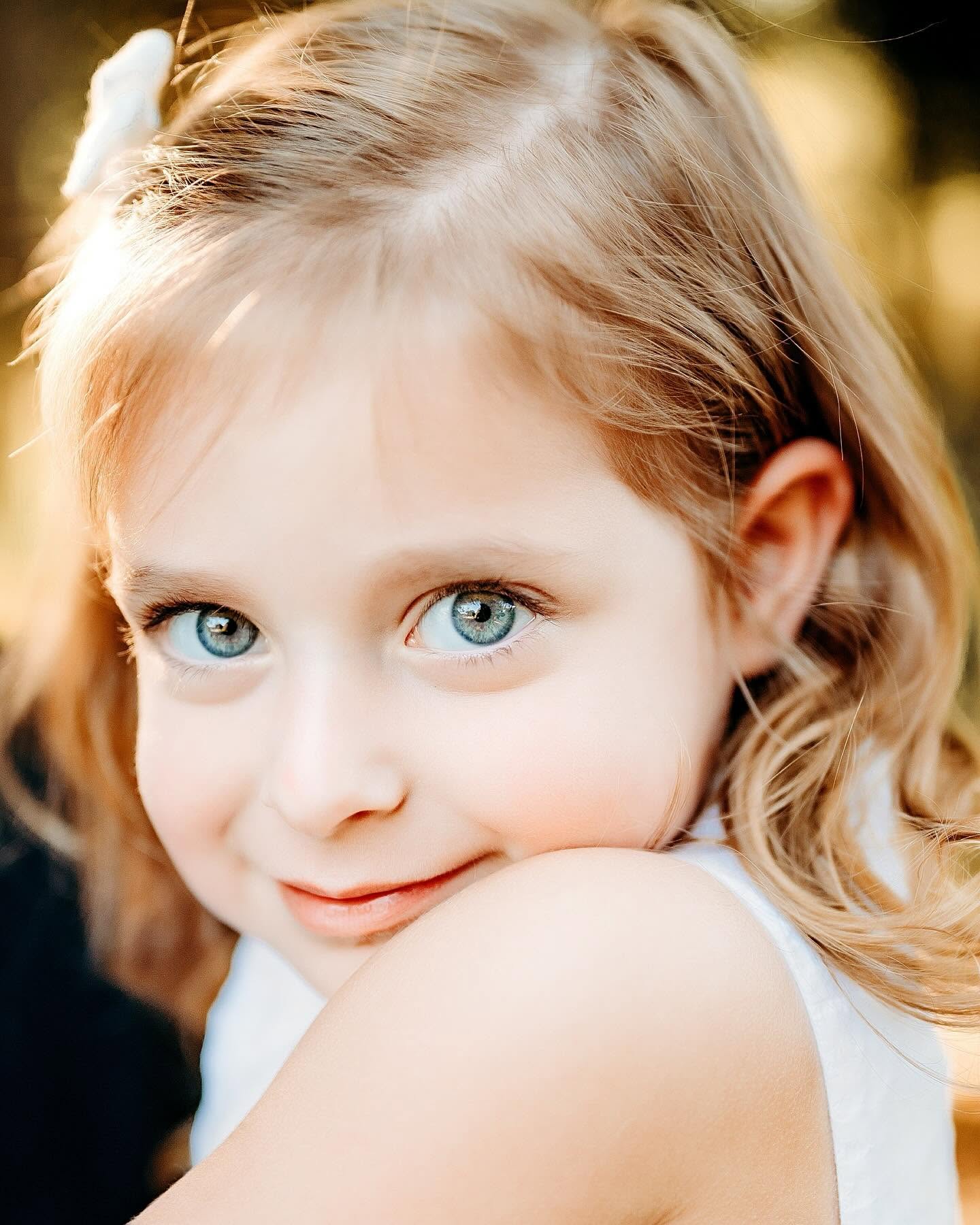 Close-up of a young girl with blue eyes and blonde hair, smiling softly, outdoors with blurred background.