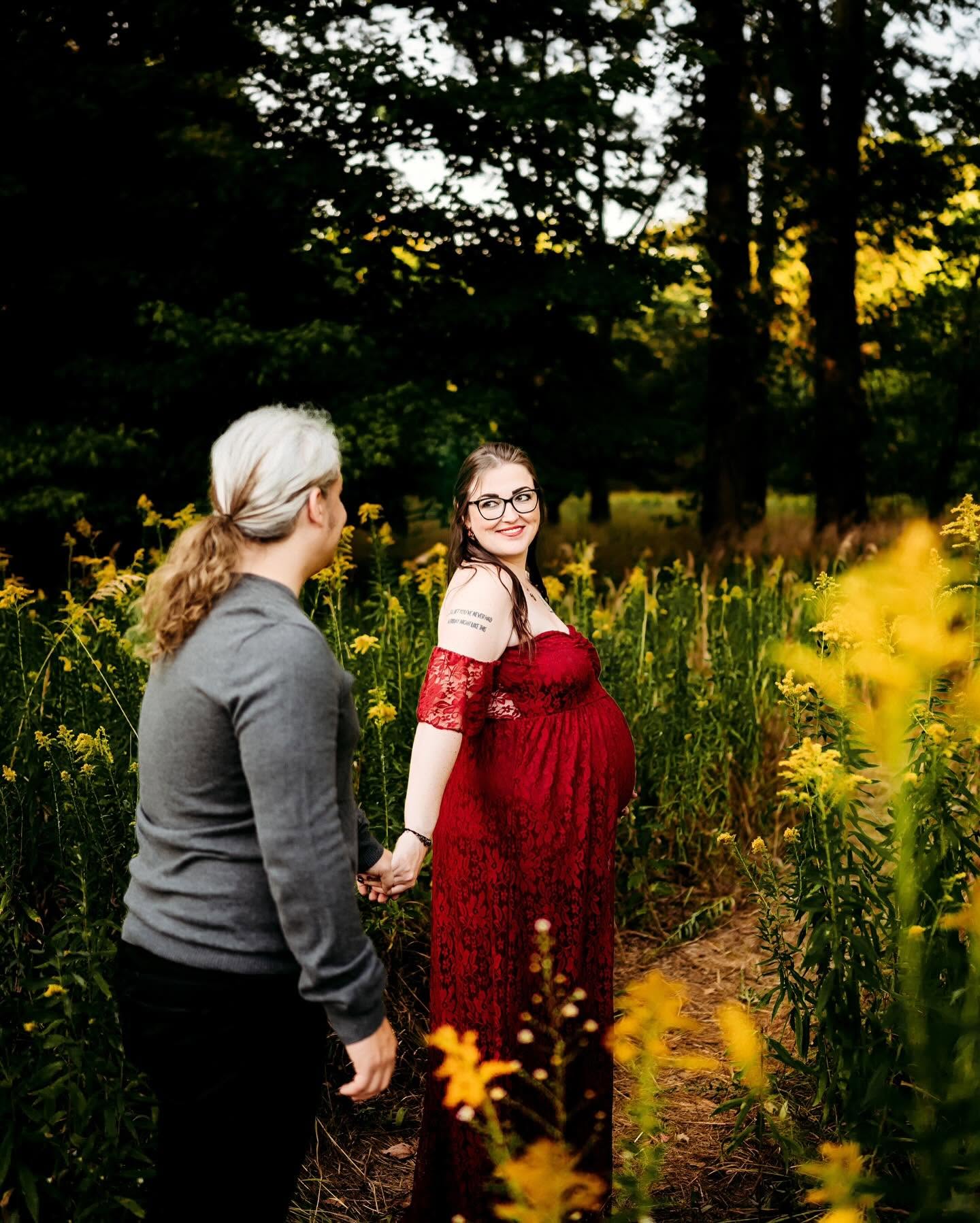 A pregnant woman in a red dress holding hands with a man in a gray sweater in a yellow flower field.