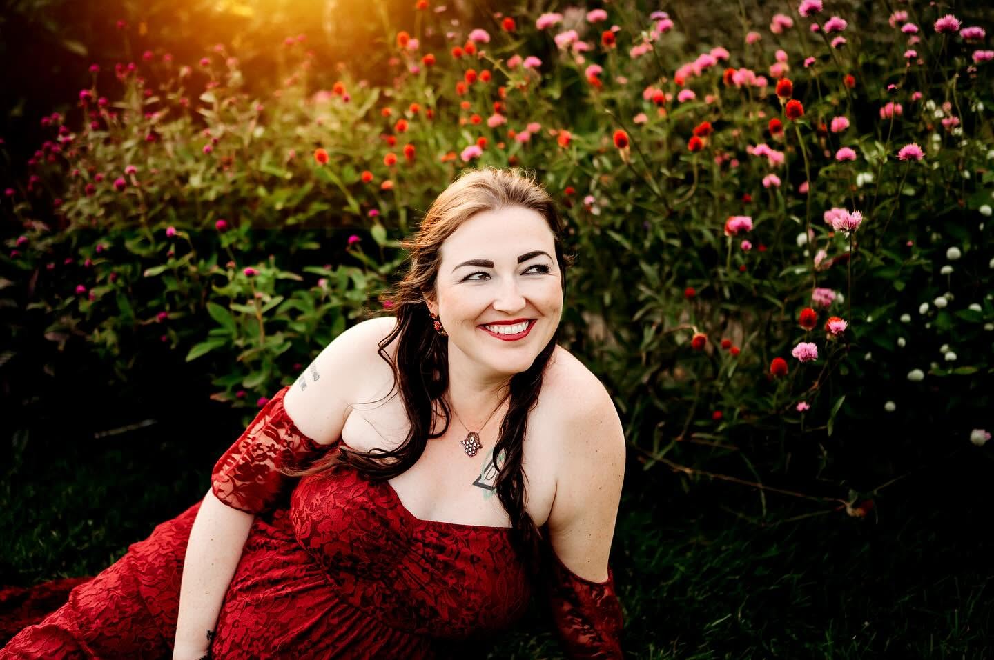 Woman with dark hair smiling in a red dress outdoors with pink flowers and greenery.