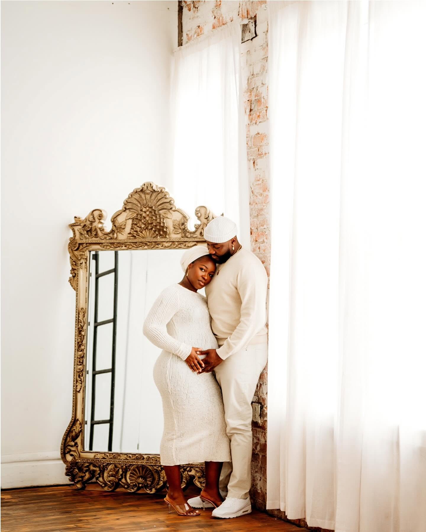 A couple stands close together in front of a large ornate mirror, holding hands, in a bright room with white curtains.