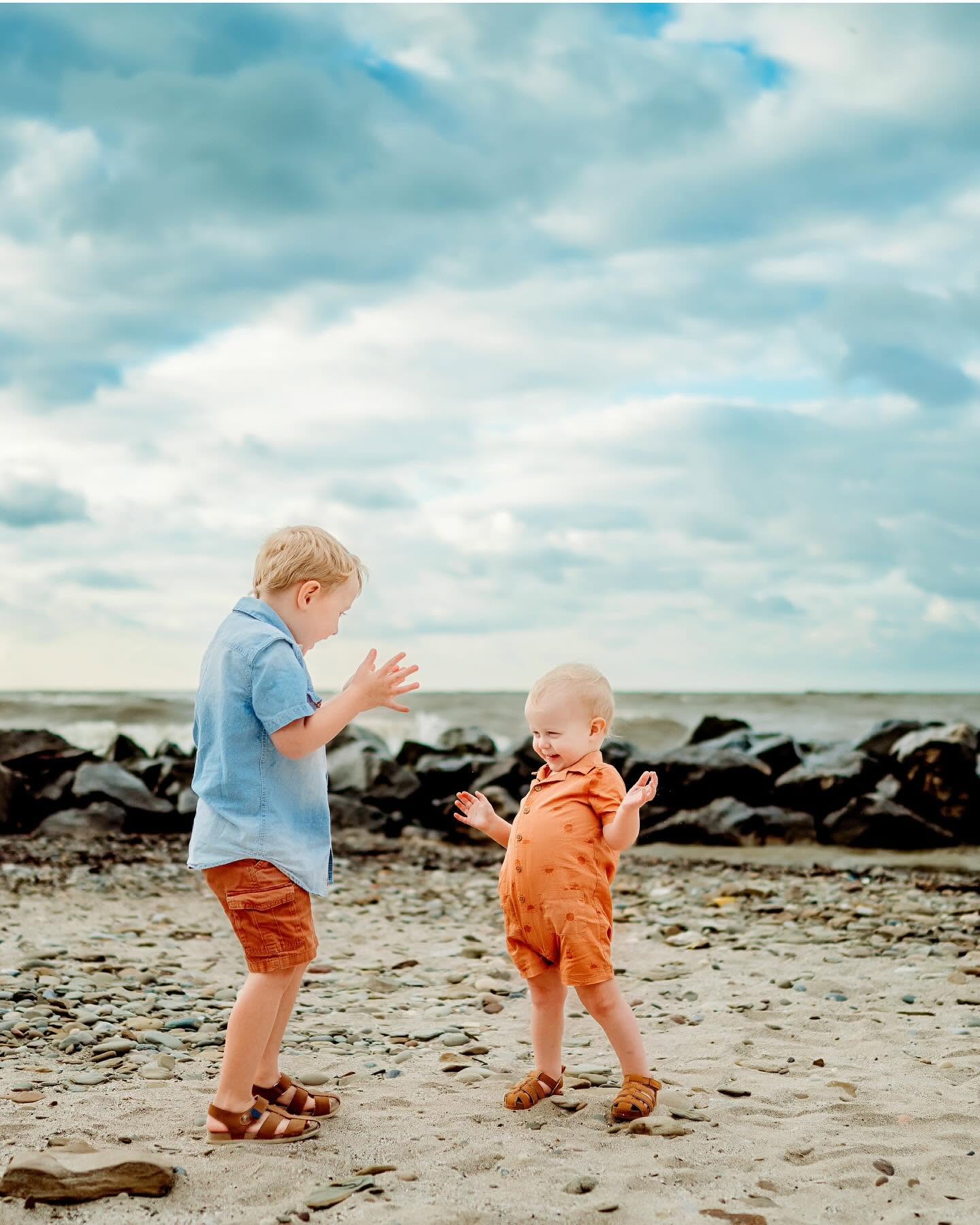Two children playing on a sandy beach with rocks and a cloudy sky in the background.