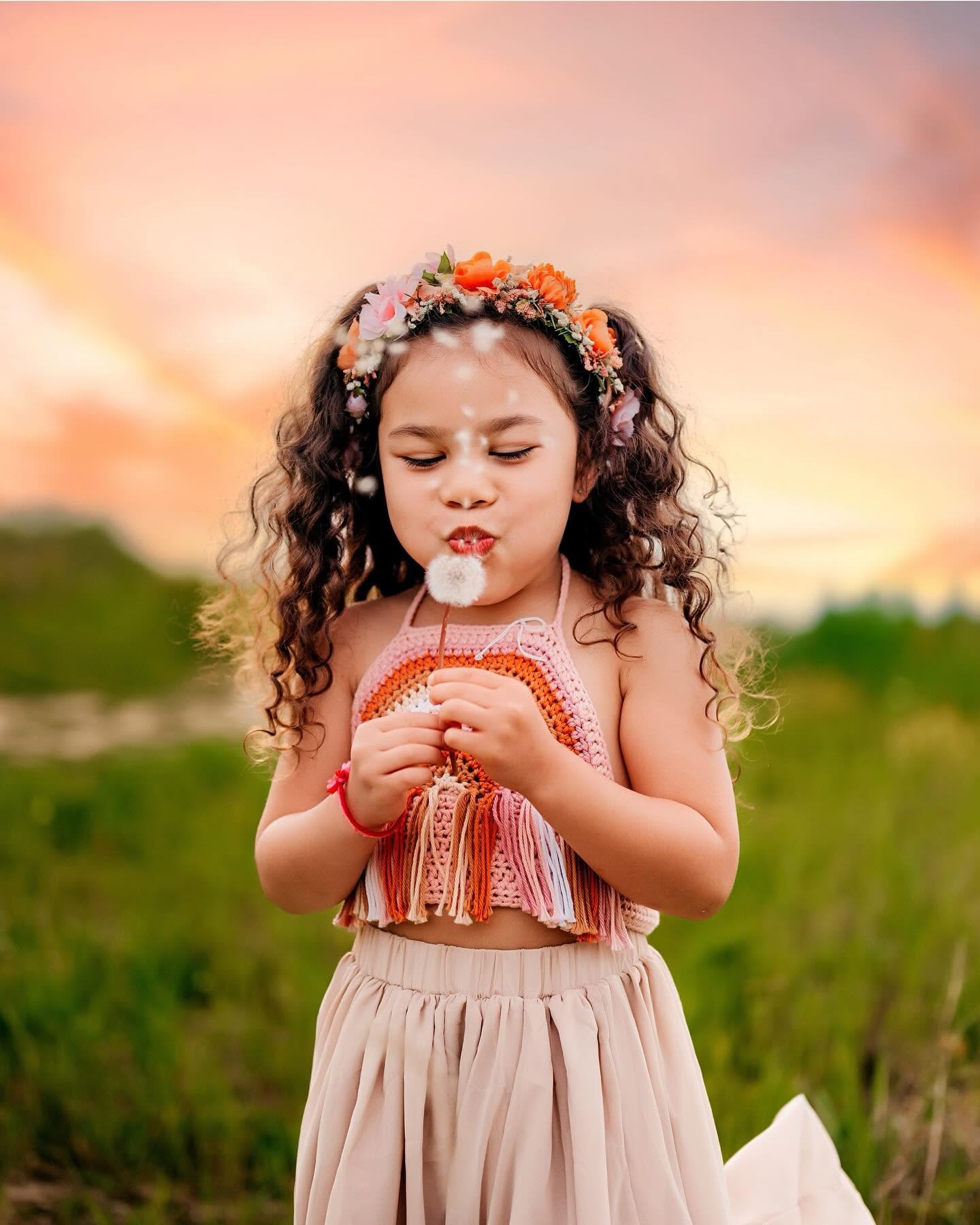 Young girl with curly hair wearing a floral headband and colorful top, holding a dandelion outdoors at sunset.
