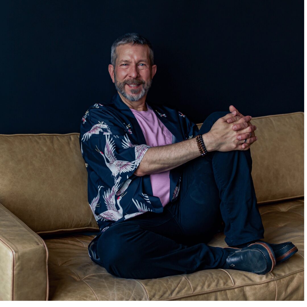 Man with gray hair and beard sitting on a brown couch, wearing a patterned jacket and pink shirt, smiling.