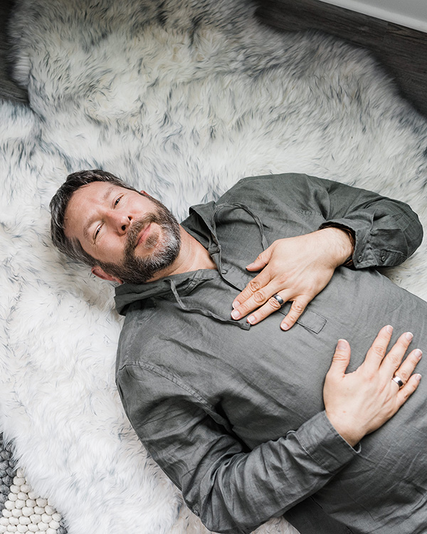 Man with beard lying on a fluffy white rug, wearing a gray jacket, with hands on chest and stomach, eyes open.