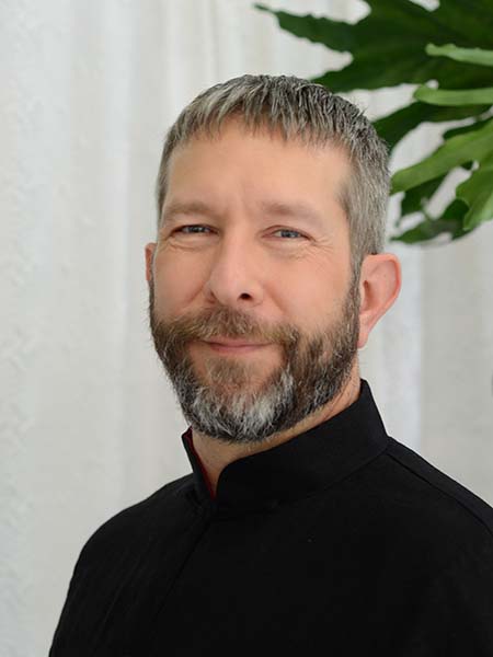 Portrait of a man with short hair and a beard, wearing a black shirt, smiling softly, with a plant in the background.