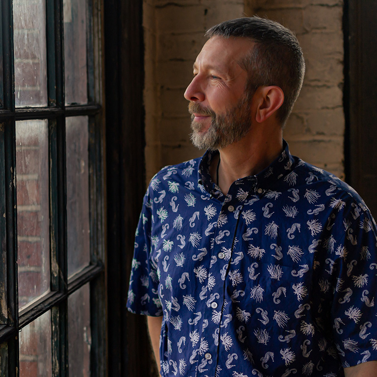 Man with short hair and beard looking out window, wearing a patterned blue shirt, in a room with brick wall.