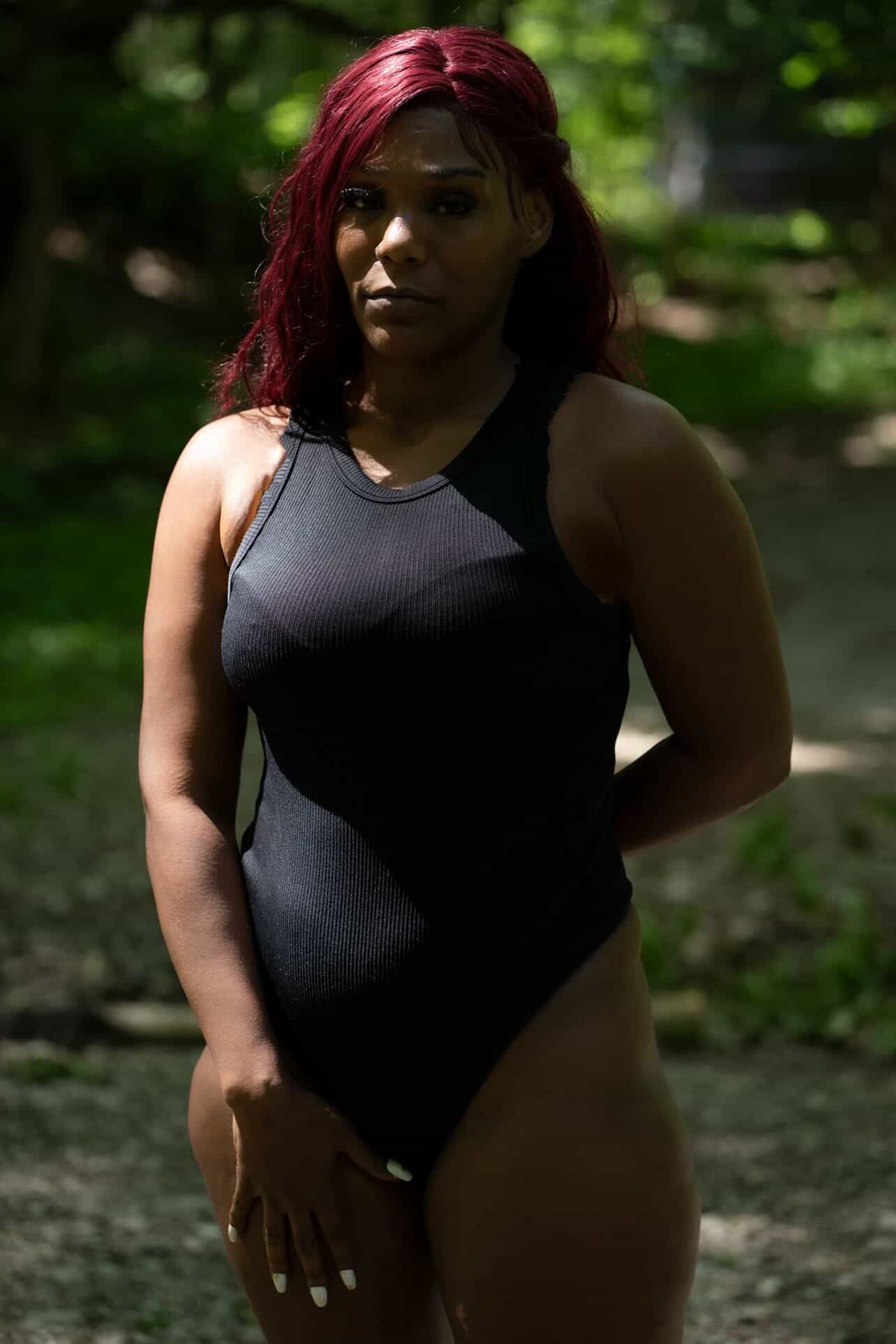 Woman with red hair standing outdoors in a black sleeveless top, with trees and sunlight in the background.