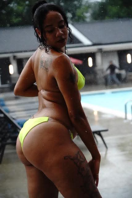 Woman in a neon yellow bikini by a poolside, wet hair, with a tattoo on her thigh, outdoors on a rainy day.