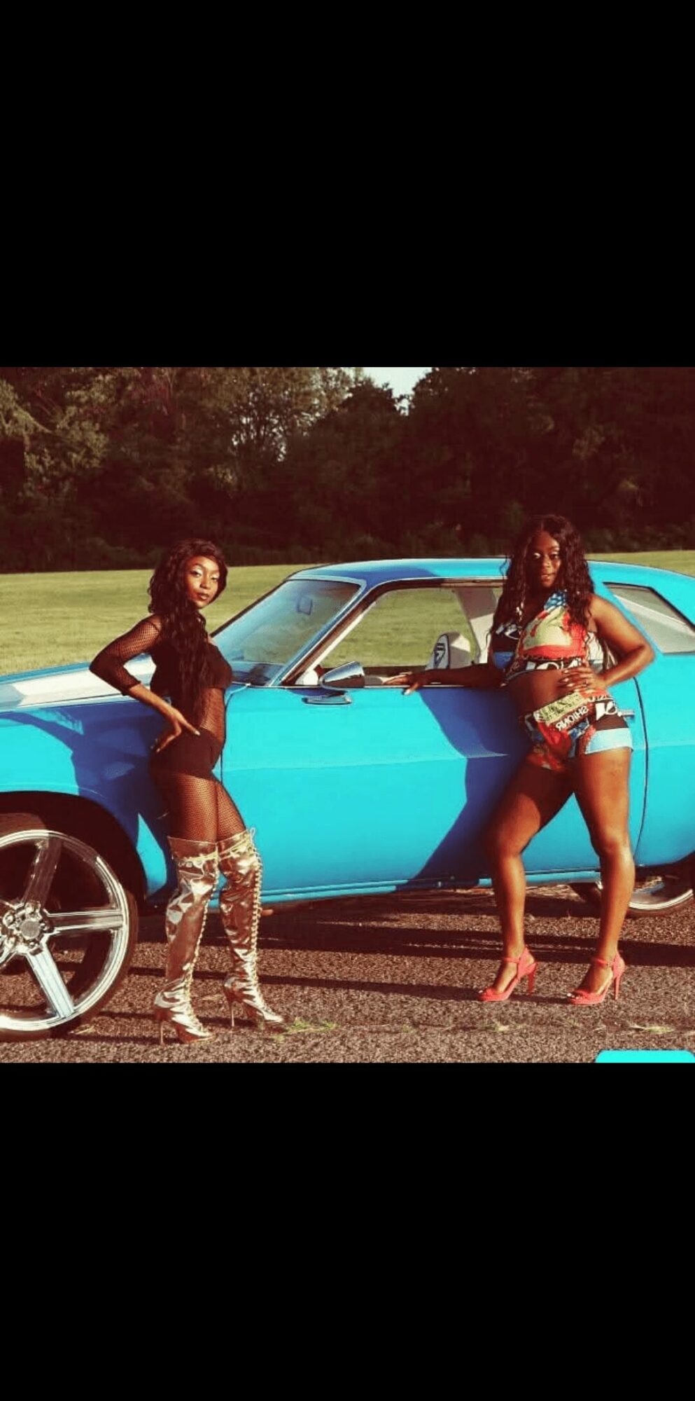 Two women in colorful outfits stand beside a blue vintage car outdoors, with trees in the background.