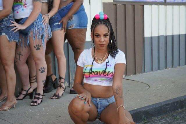 Young woman crouching on sidewalk with group of women in background, wearing a white crop top and denim shorts.