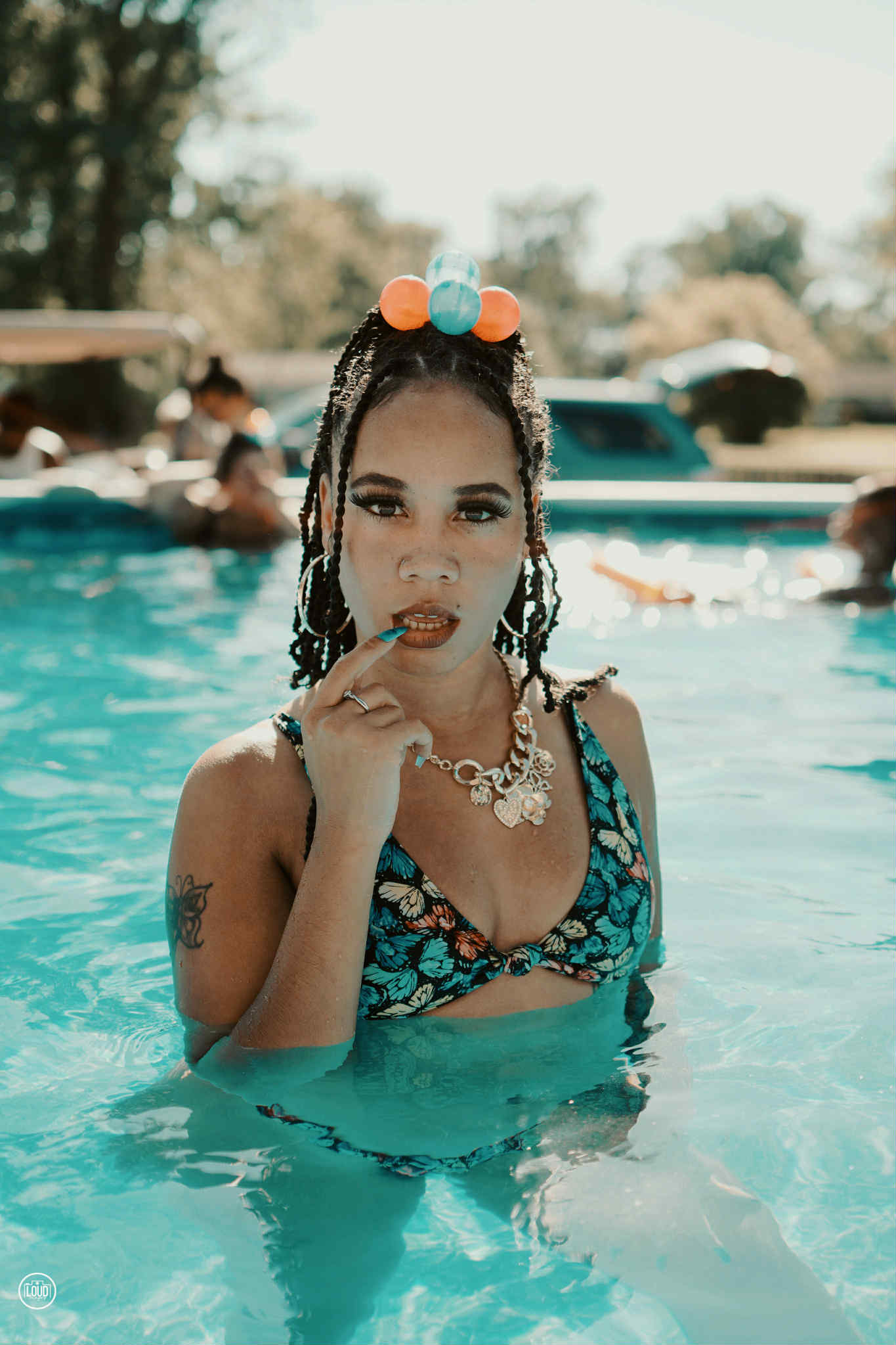 Young woman in a swimsuit with jewelry, standing in a swimming pool, holding her chin, with people and trees in background.