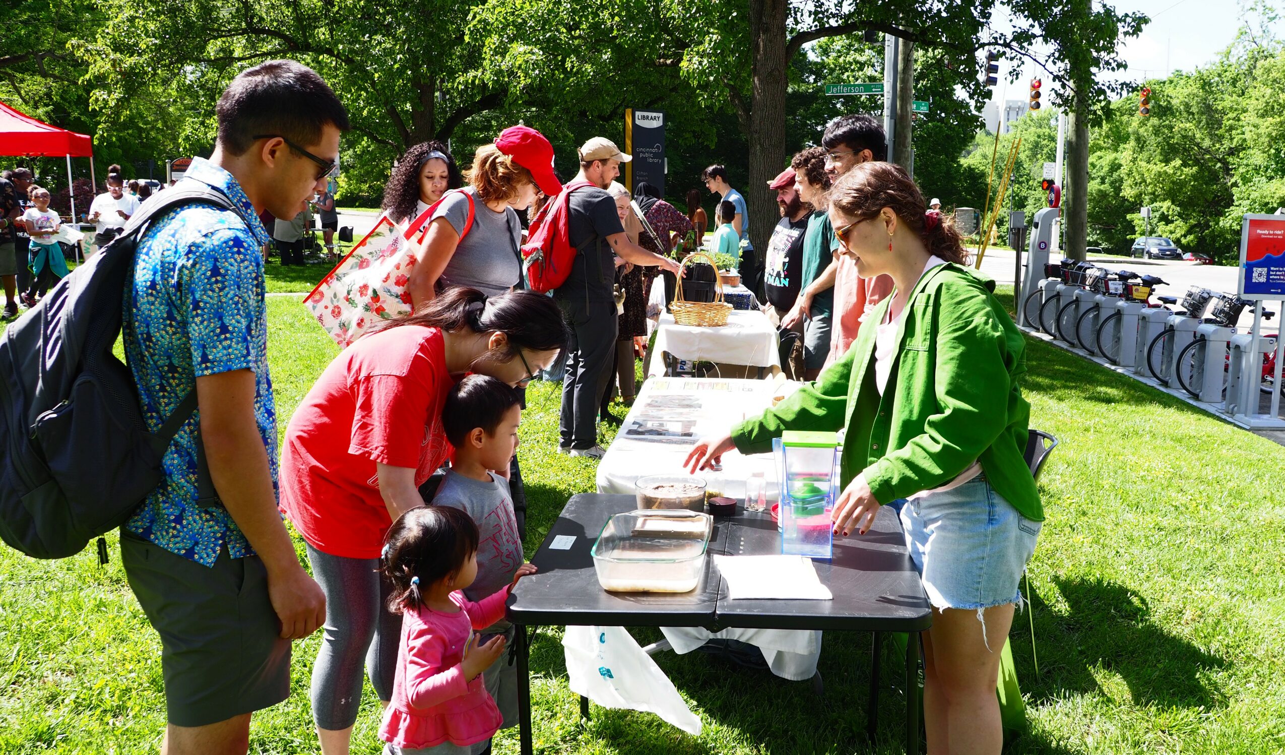People gather around tables outdoors on a sunny day, with trees and bicycles in the background.