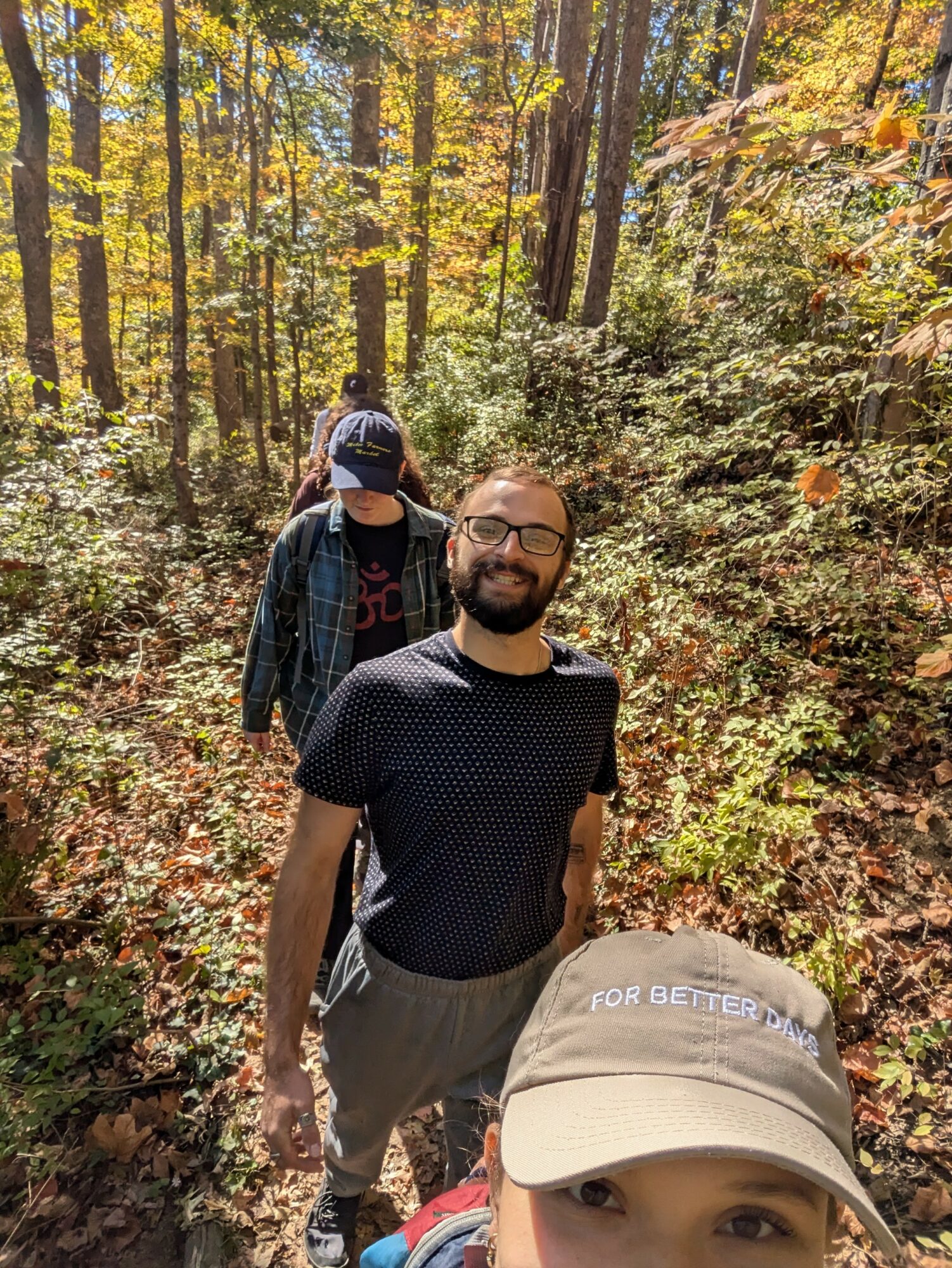 Two people walking in a forest with trees and sunlight, one smiling at the camera wearing a cap, the other wearing a mask and glasses behind.