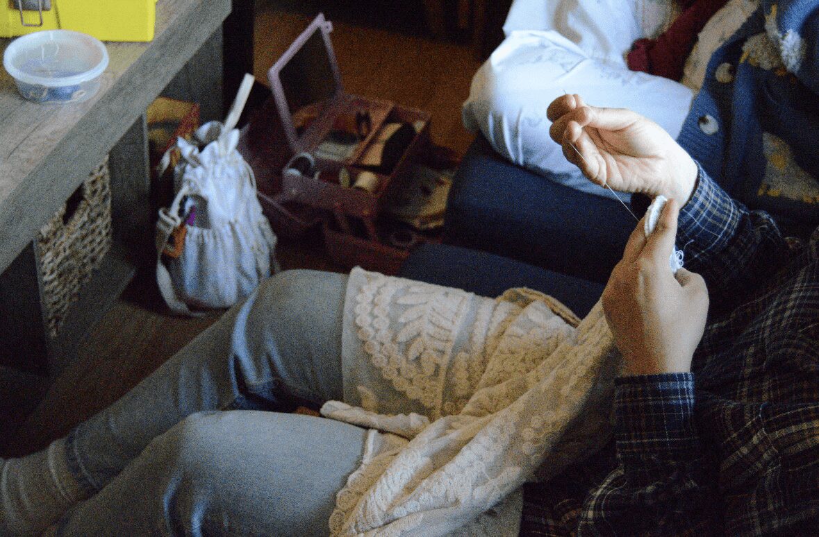 Two people sitting on a couch, one with hands raised, a table with a basket and a mirror nearby, and a person in white pants.