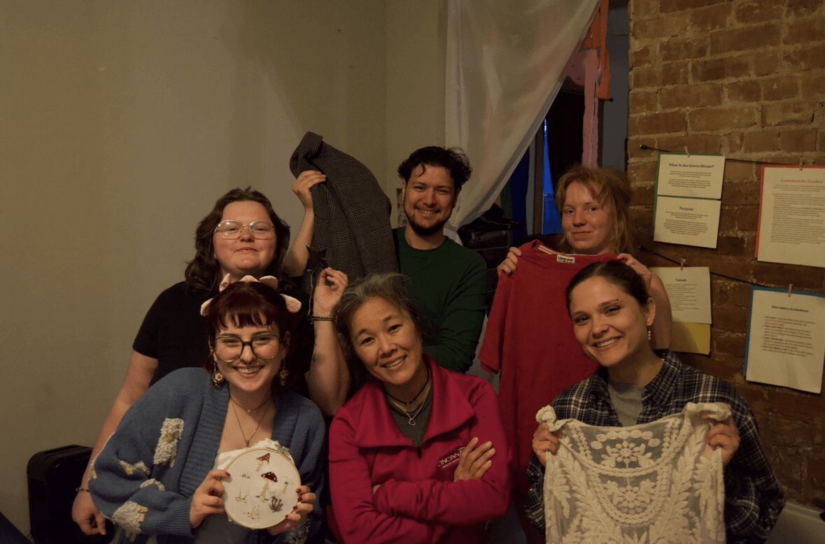 Group of six people smiling indoors, some holding clothing or fabric, with a brick wall and notices in the background.