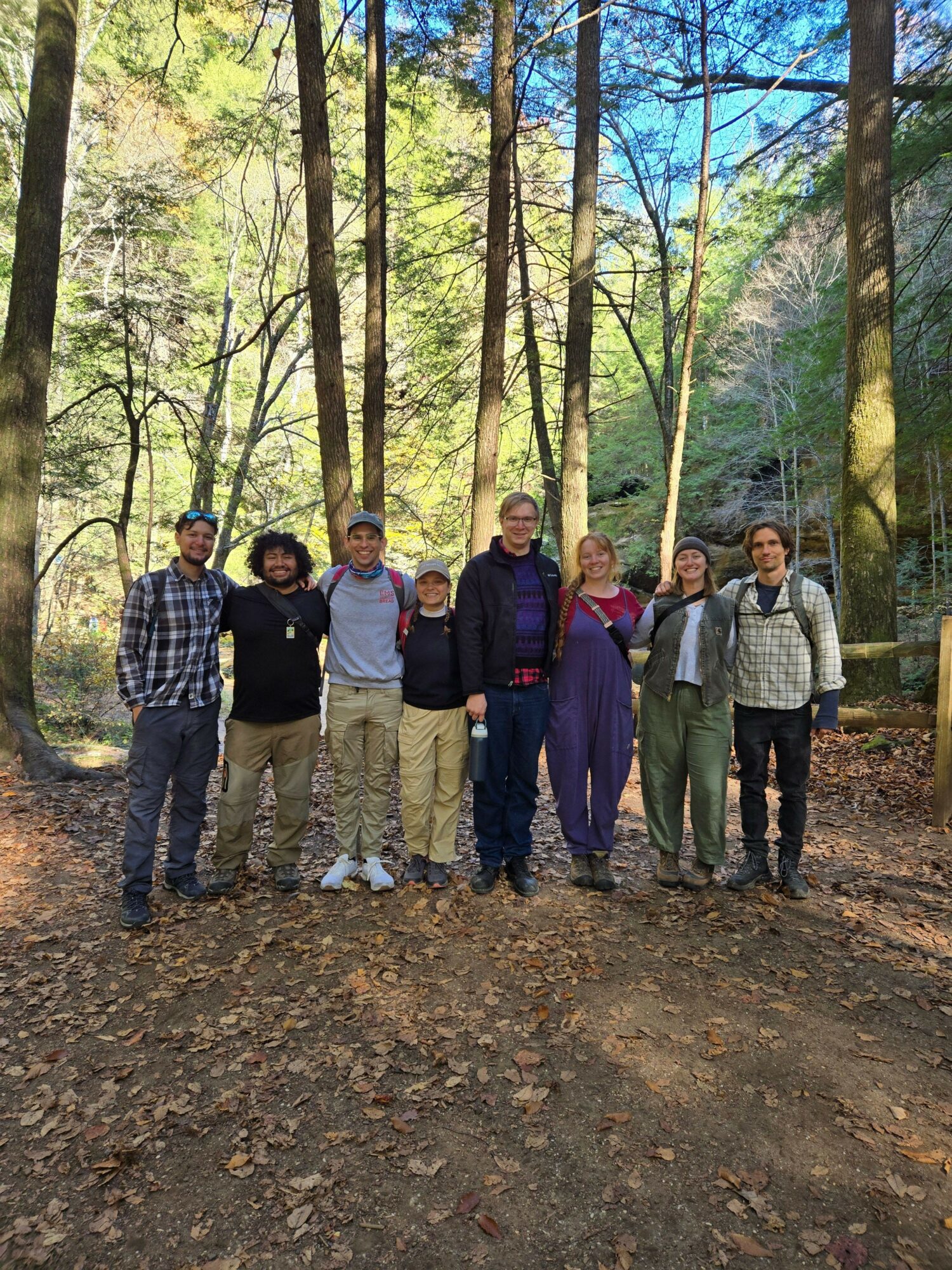 Group of six people standing on a dirt path in a forest with trees and sunlight, posing for a photo.