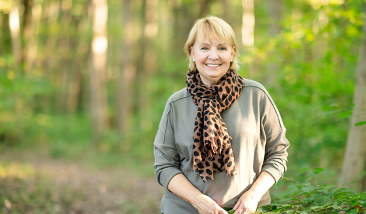 Smiling woman with short blonde hair wearing a leopard print scarf and gray jacket outdoors in a green forest
