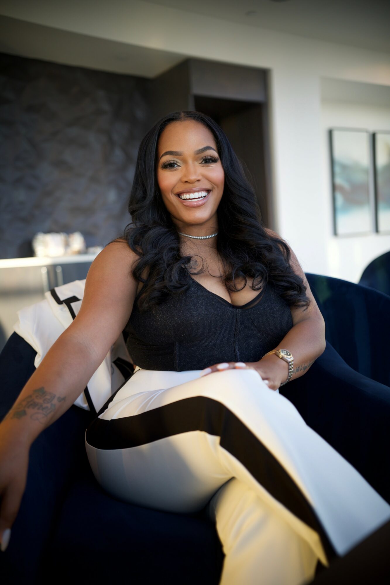 Smiling woman with long dark hair, wearing a black top and striped pants, sitting indoors with a modern background.
